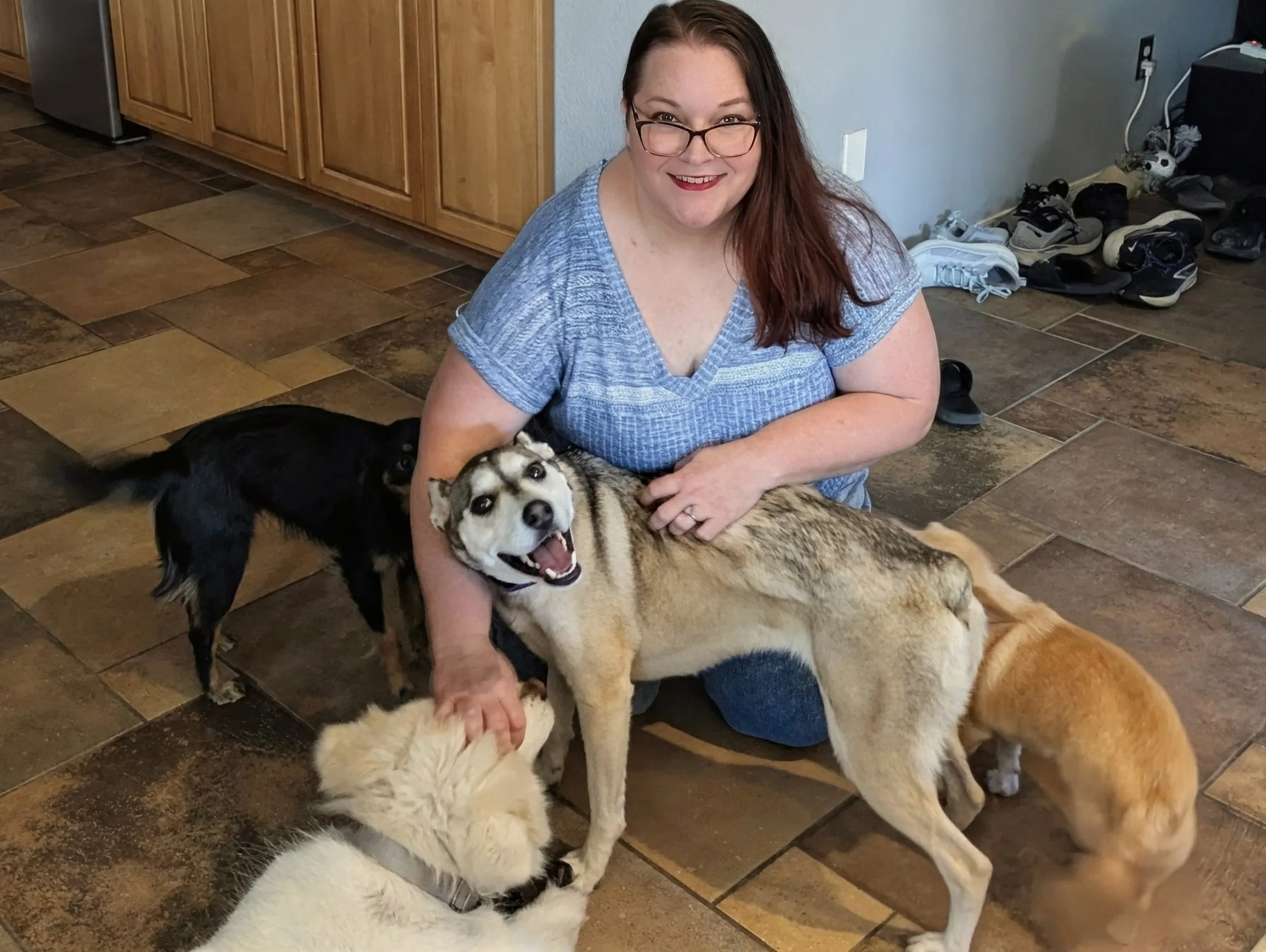 A woman with long brown hair, glasses, and red lipstick smiling while sitting on a tile floor with cute puppies and dogs around her. In the background, there are wooden cabinets and a pile of shoes near the wall.