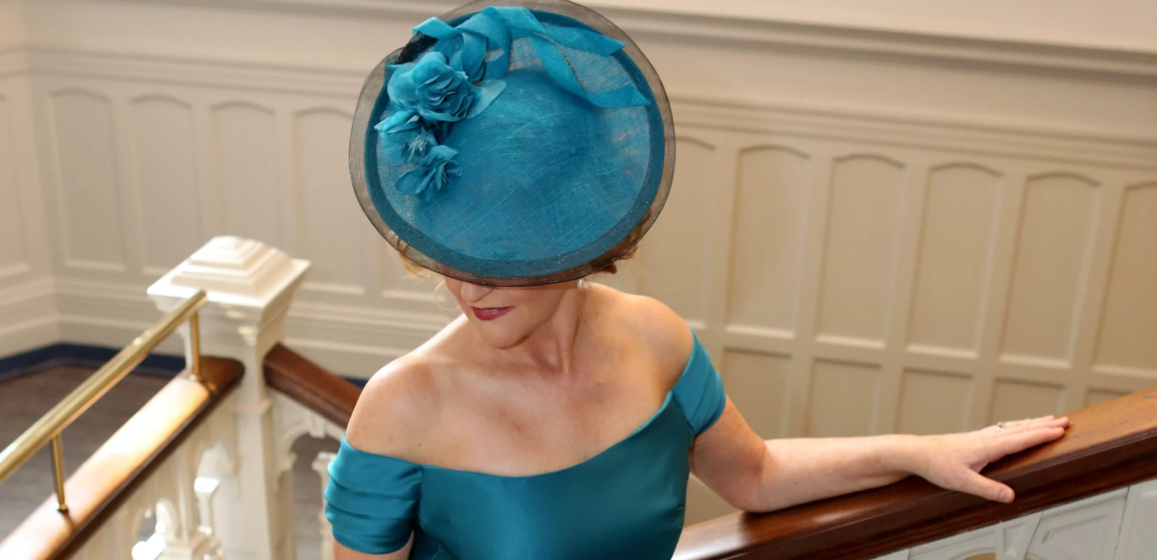 Model wearing a teal dress and a large teal John Paul Millinery hat with fabric flowers, standing on a staircase at Bishops Gate Hotel, Derry.