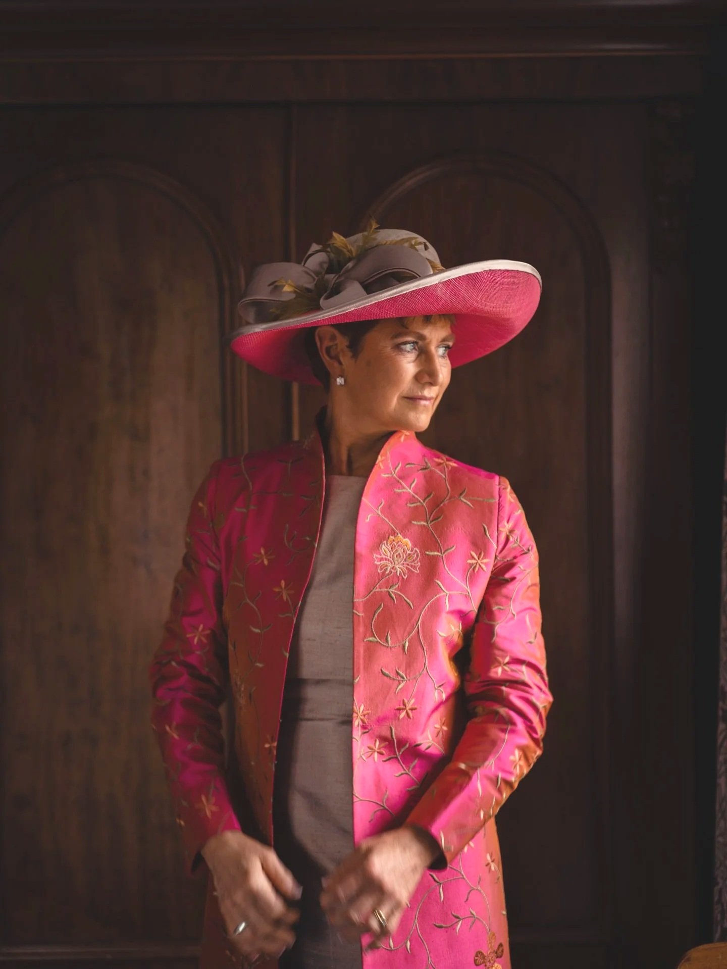 A woman in a large pink John Paul Millinery hat and a pink embroidered jacket standing in front of a wooden cabinet, looking to her right.