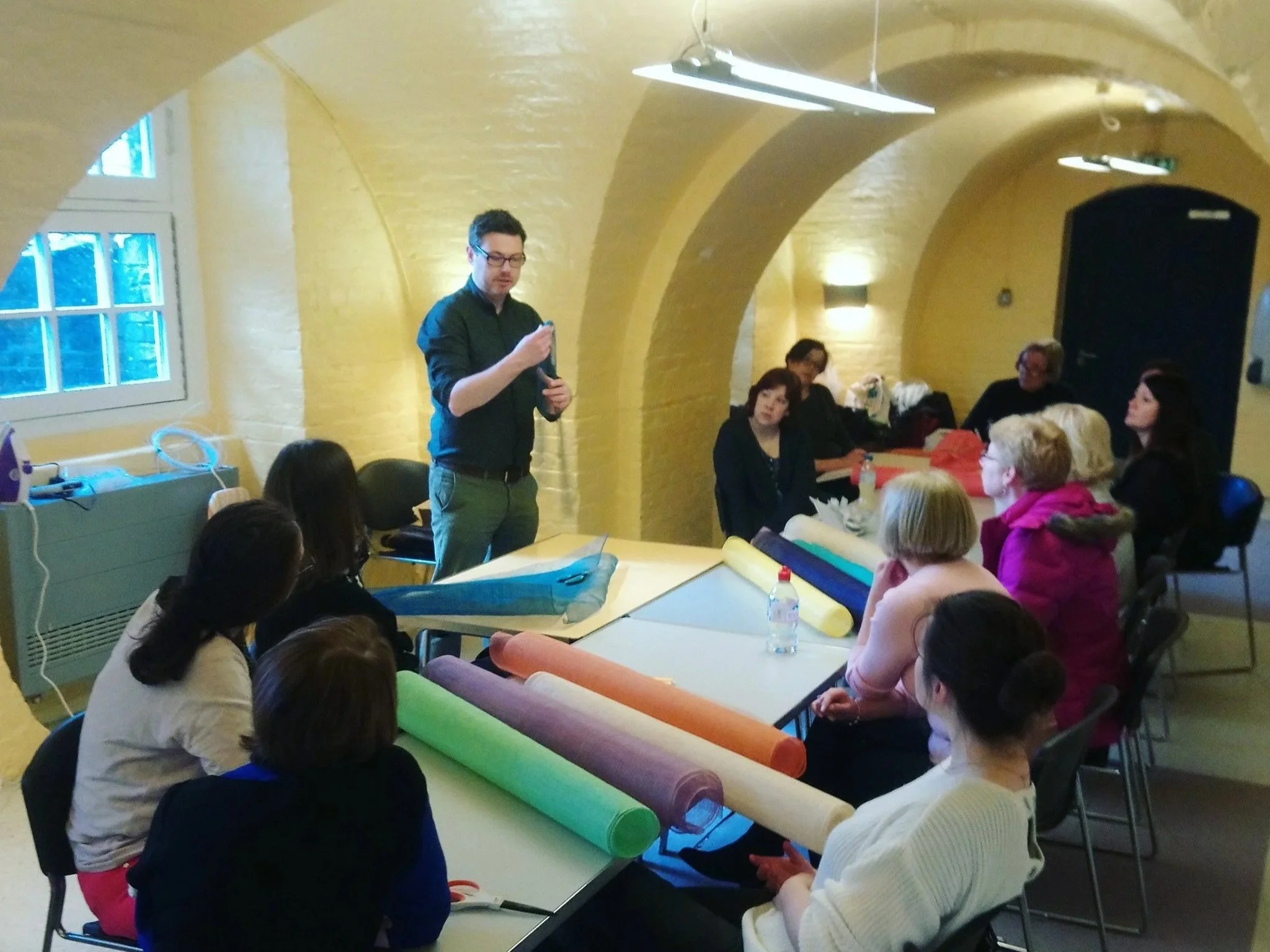 A man stands at the head of a table giving a presentation to a group of women seated around the table. The table has rolls of colored paper and tools, suggesting a workshop setting in a room with yellow brick walls and arched ceilings.