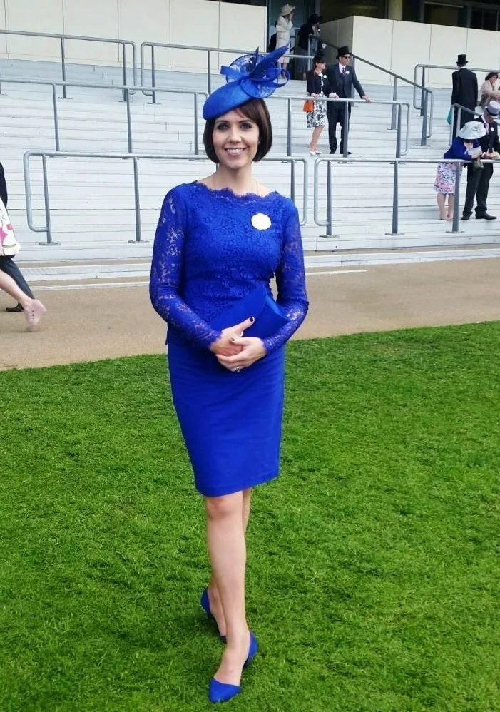 Woman dressed in a royal blue lace dress and matching heels, wearing an elaborate John Paul Millinery blue fascinator, standing at Royal Ascot.