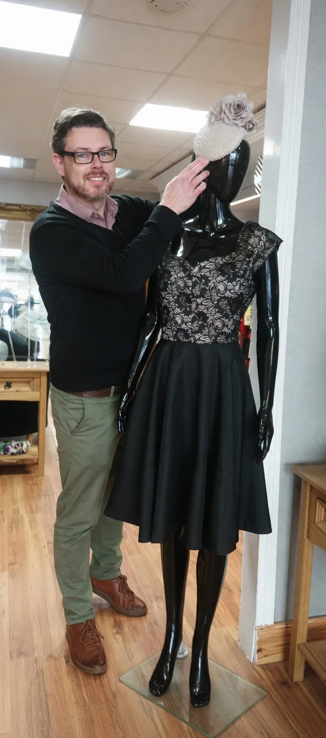 John Paul Deehan of John Paul Millinery, standing next to a mannequin dressed in a black finished with John Paul Millinery hat with a decorative flower.