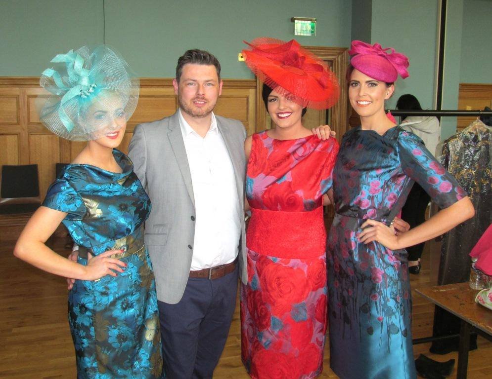 Four people are posing together in a room, three women in colorful floral dresses with large, elaborate hats and one man in a gray blazer and white shirt. The women are wearing vibrant, patterned dresses and fancy hats, while the man is dressed more casually. The room has wooden paneling on the walls and some clothing racks in the background.