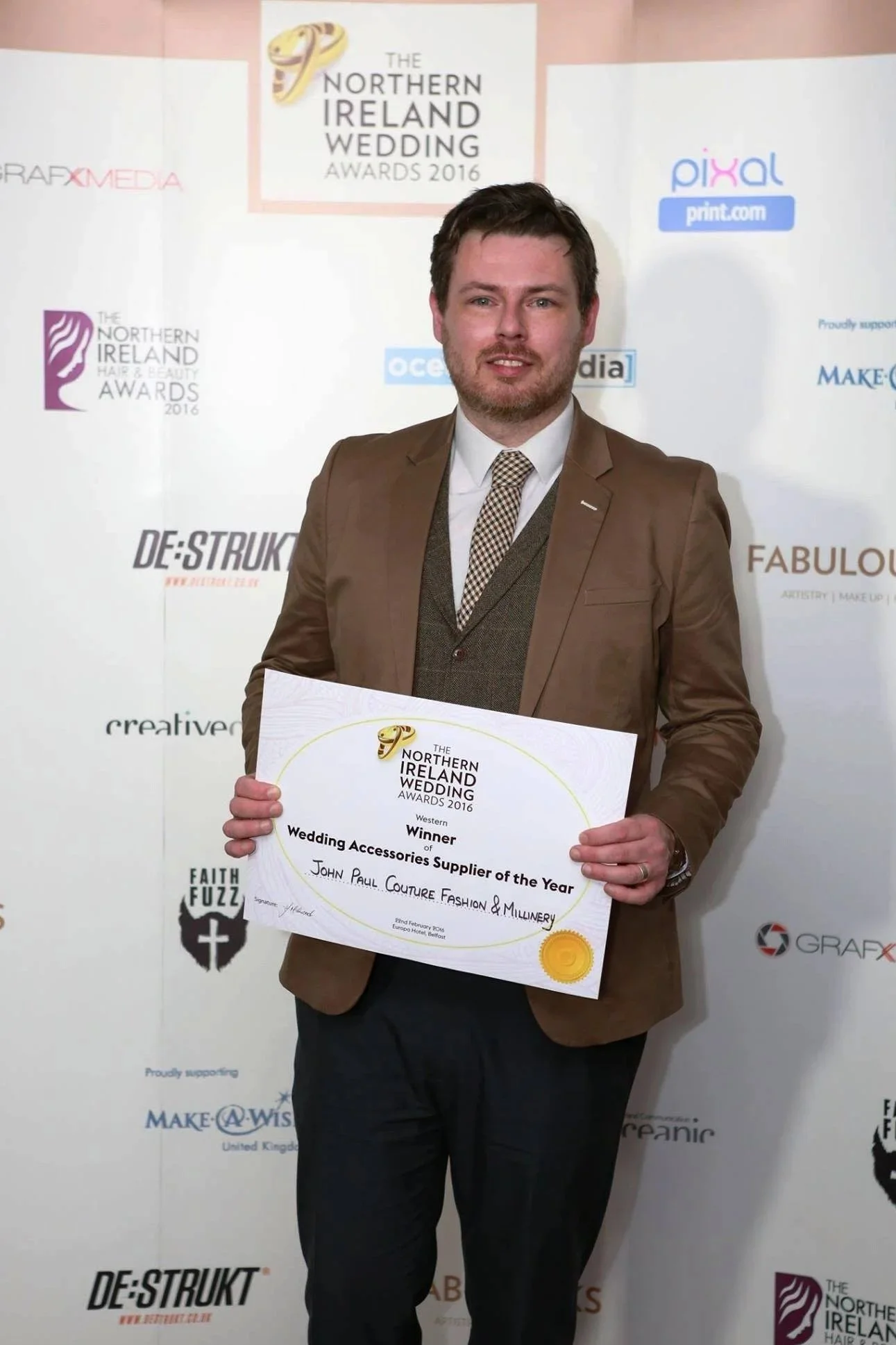 A man in a brown suit holding a certificate at the 2016 Northern Ireland Wedding Awards, standing in front of a backdrop with various sponsor logos.