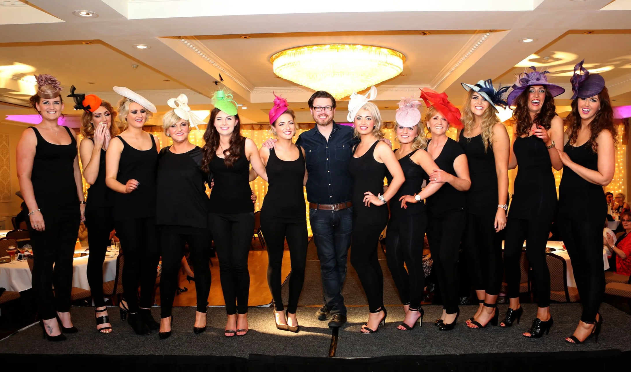 Group of women and one man standing together on a stage at a hat-themed event, all wearing black clothing and colorful fancy hats, indoors with a large chandelier overhead.