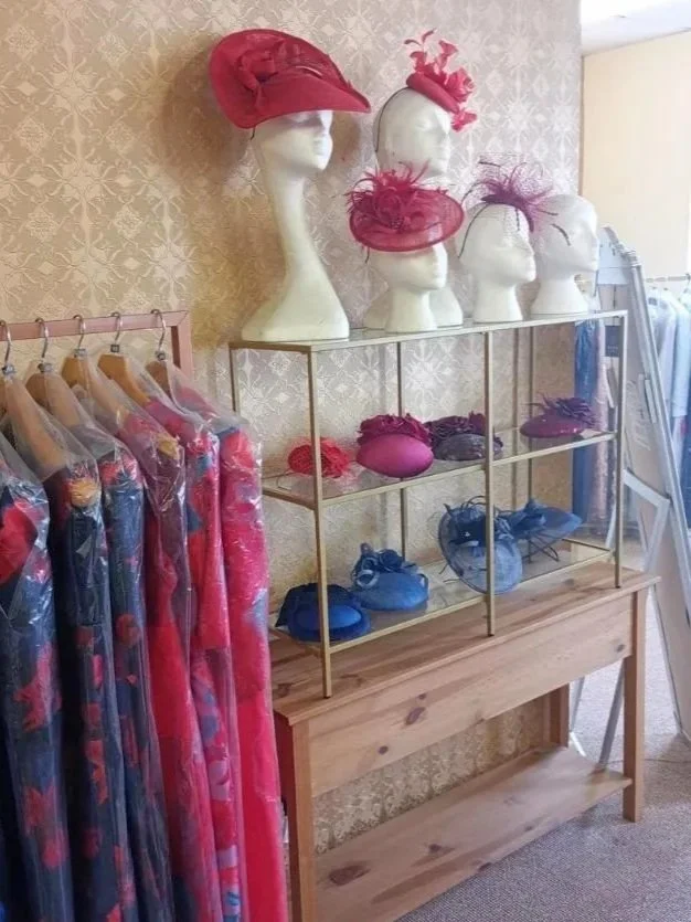 Display of pink and blue hats and headpieces on mannequins, with colorful dresses hanging nearby.