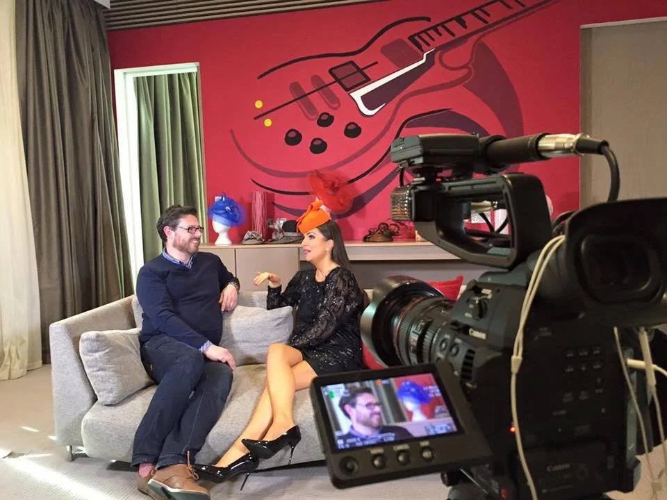 Two people sitting on a beige couch in front of a pink wall with a guitar mural, being filmed by a professional camera, with a woman talking animatedly and a man listening and smiling.