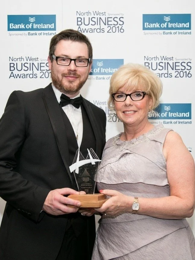 A man in a tuxedo and a woman in a light gray dress standing together at the North West Business Awards 2016, holding an award trophy between them, with a backdrop displaying the event logo and Bank of Ireland sponsorship.