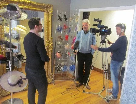 An interview being conducted in a store filled with hats and accessories. A man with gray hair and a blue jacket speaks into a microphone held by a cameraman. Another man with glasses, wearing a black sweater, listens. A professional camera on a tripod records the interview.