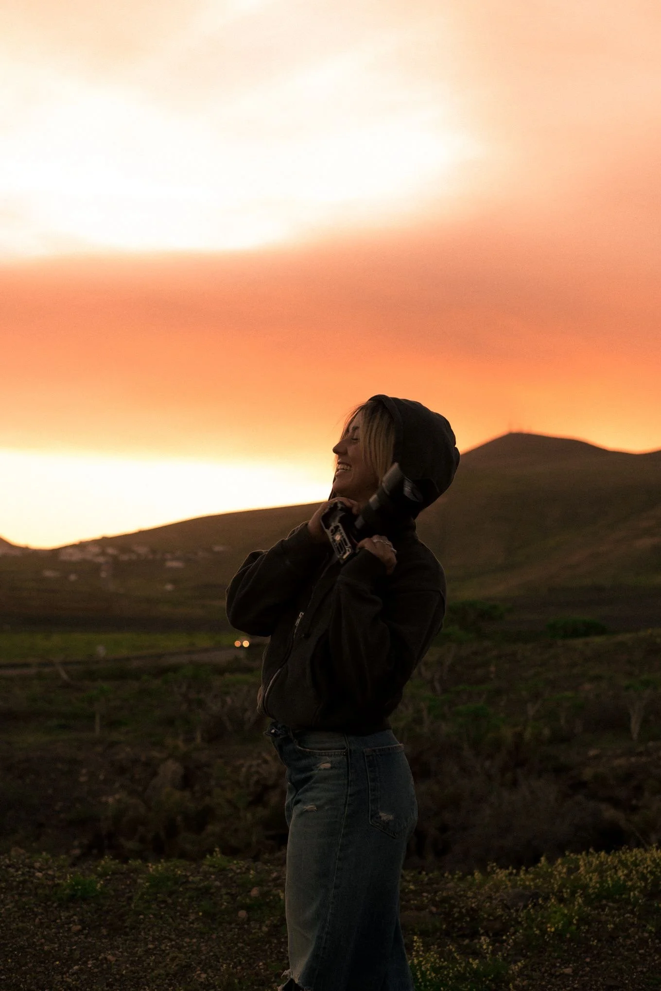 Una donna sorridente con cappuccio, indossa una giacca e jeans, si trova all'aperto durante il tramonto, con montagne sullo sfondo e un cielo dai toni caldi.