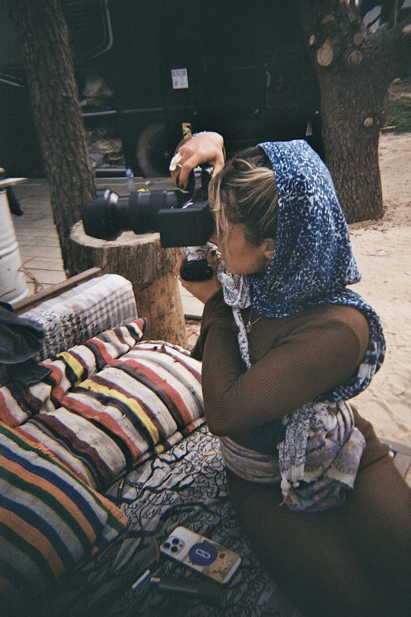 Una persona indossa una sciarpa blu con motivo bianco, è seduta o in piedi vicino a un letto con coperte colorate a righe. La persona sta guardando attraverso un binocolo rivolto verso un letto. Sul pavimento ci sono vari oggetti tra cui un mazzo di carte e un videogioco.