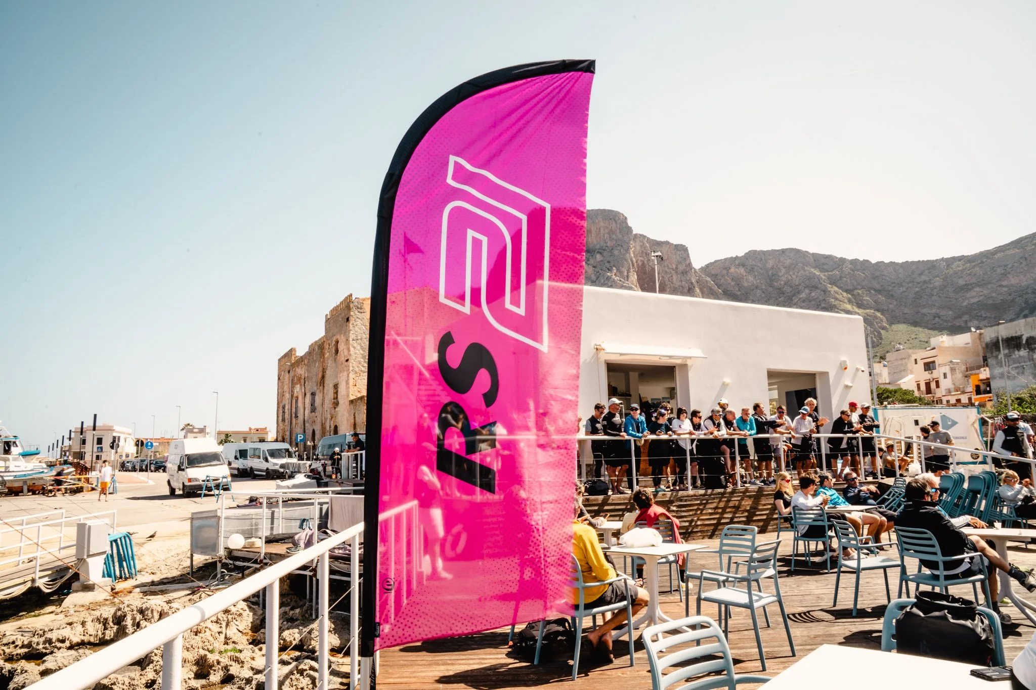 Gruppo di persone sedute e in attesa di un evento all'aperto, con bandiera rosa, sul lungomare di una città, sullo sfondo montagne e edifici