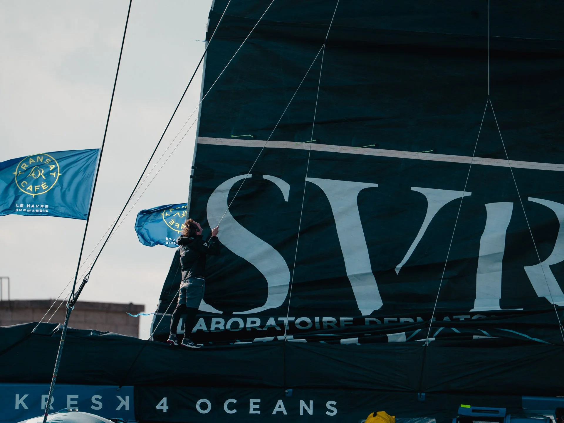 Un ragazzo che si trova su una barca, pare in fase di preparazione per una regata, che issa una grande vela nera con le parole 'KRES 40 OCEANS' e tiene in mano una bandiera blu con un logo circolare e il testo 'TRANSAT CAFE LE HAVRE NORMANDIE'.