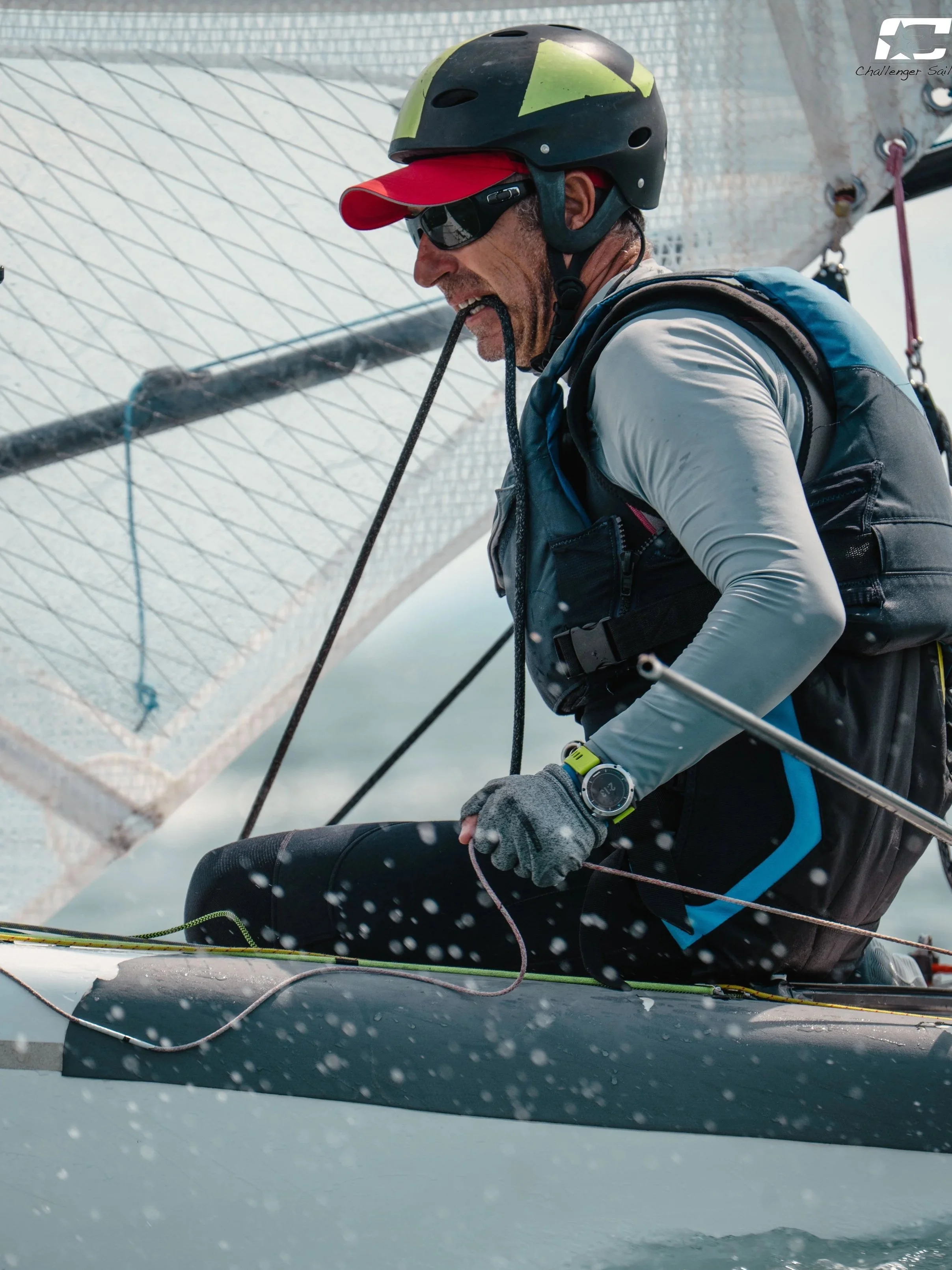 Velista mentre si prepara a salire a bordo di una barca a vela, indossa casco, occhiali da sole e abbigliamento tecnico, con vele e attrezzatura visibile.