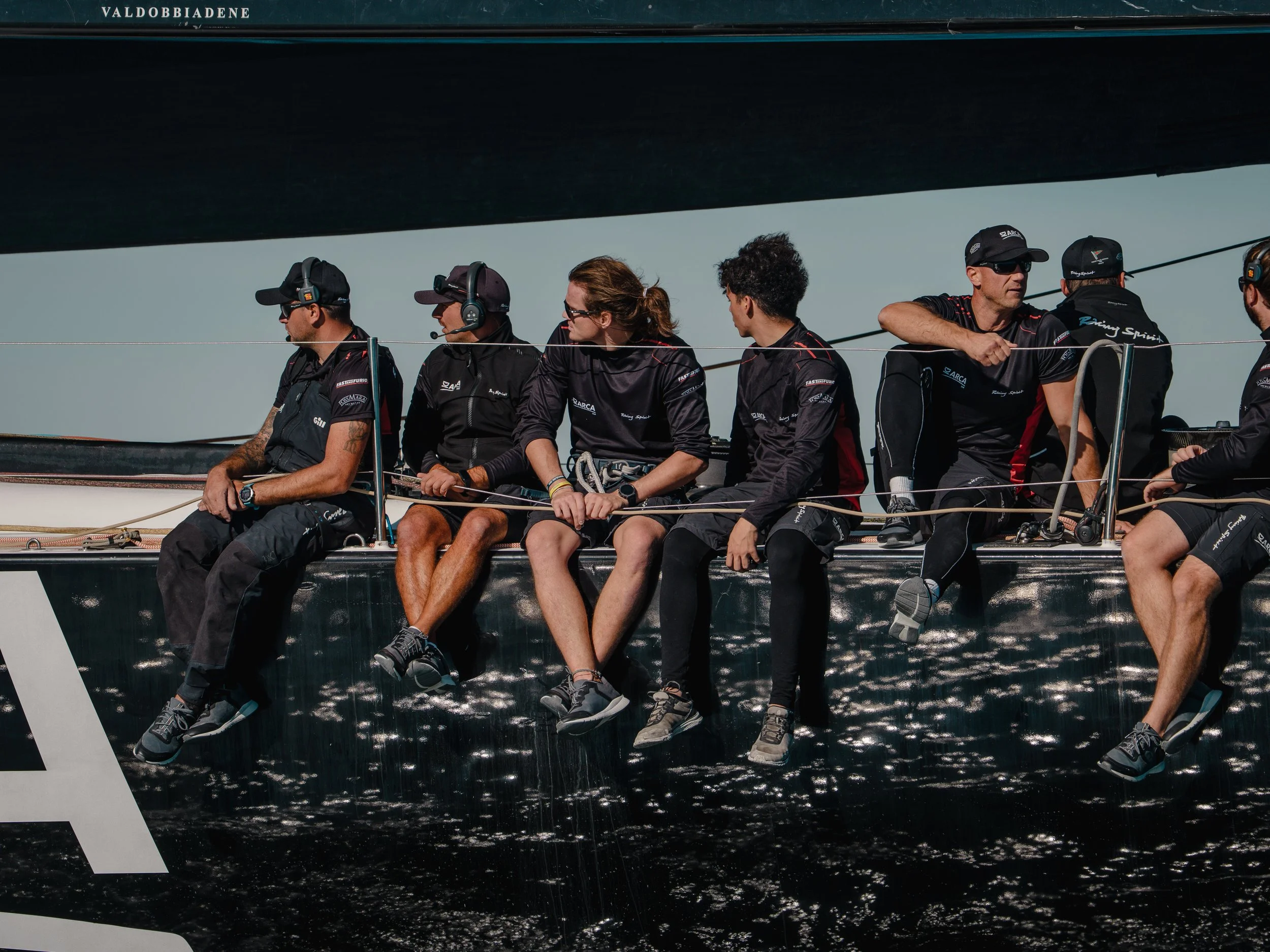 Foto di un gruppo di persone sedute su una barca a vela, indossando caschi e abbigliamento sportivo, che appaiono concentrati mentre si preparano durante una giornata di vela.