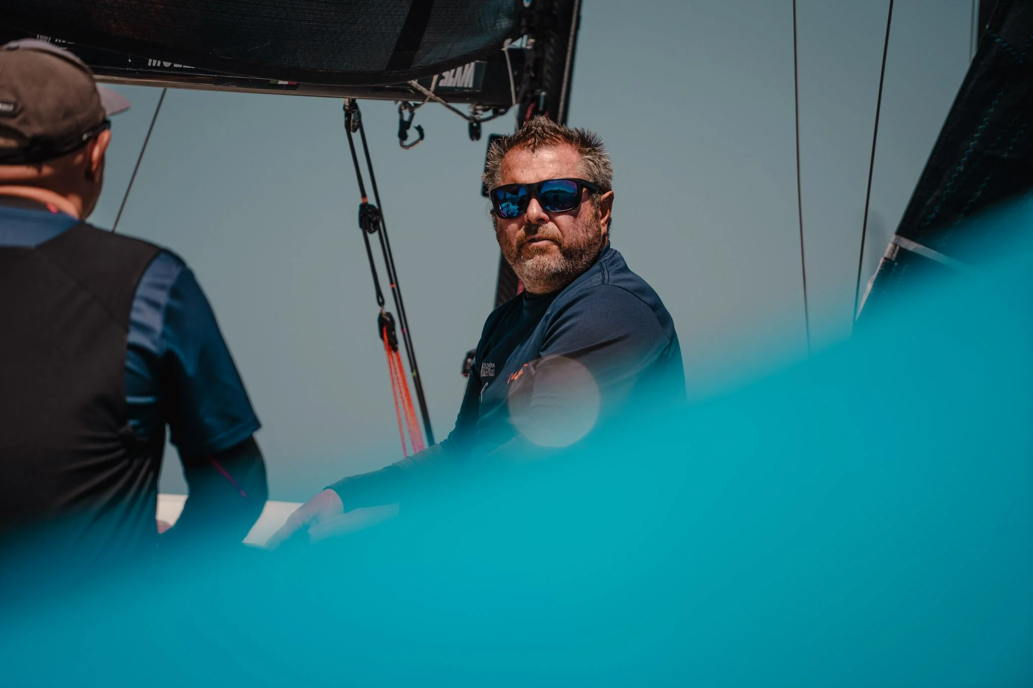 Uomo con barba e occhiali da sole seduto su una vela di barca, con persone intorno, sotto un cielo chiaro.