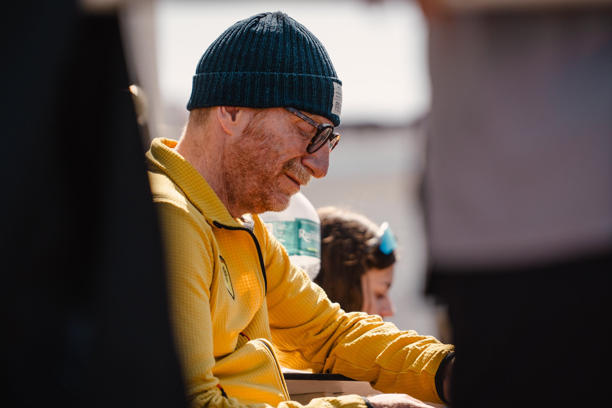Uomo con cappello invernale, occhiali e giacca gialla sorridente, seduto e che interagisce con altre persone in un ambiente esterno.
