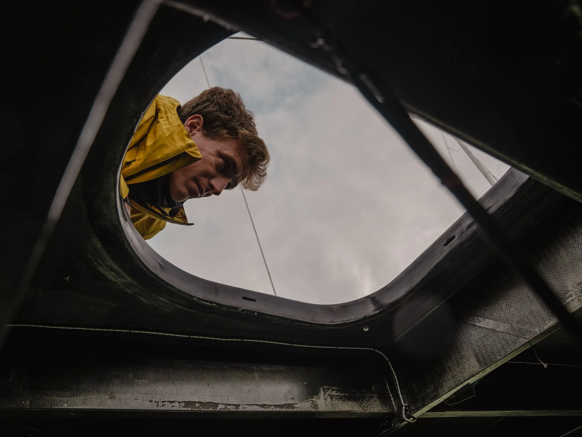 Persona con capelli castani, abbigliamento giallo, vista attraverso un foro in una struttura nera, forse parte di un veicolo o di un macchinario, con cielo nuvoloso sullo sfondo.