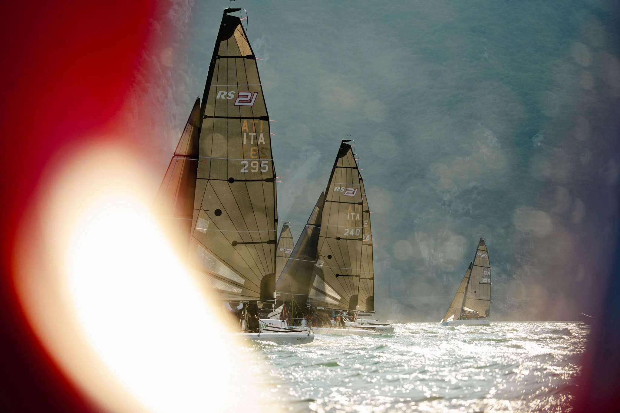 Immagine di barche a vela che gareggiano in mare sotto un cielo con nuvole, con un effetto di luce solare e un'iferografia vicino alla telecamera.