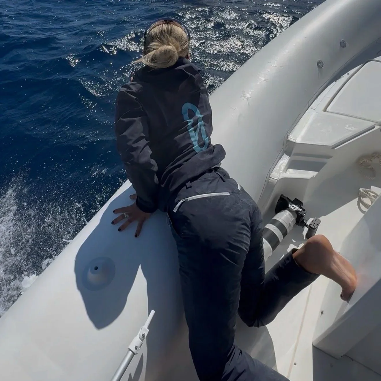 Una persona che si trova su una barca in mare aperto, appoggiata sullo scafo e con una fotocamera con grande obiettivo posizionata vicino a lei.