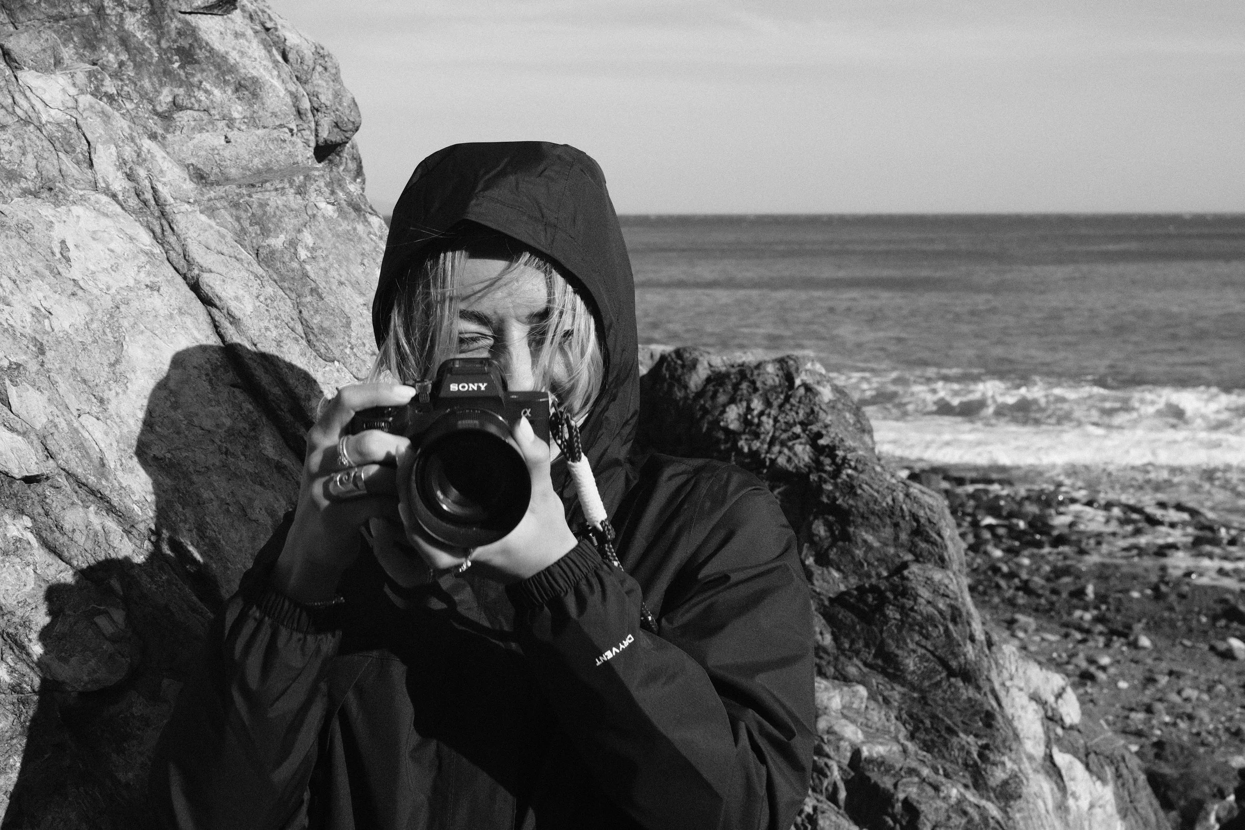 Una persona con cappuccio che scatta una foto con una fotocamera di fronte a rocce e al mare, in un paesaggio costiero.