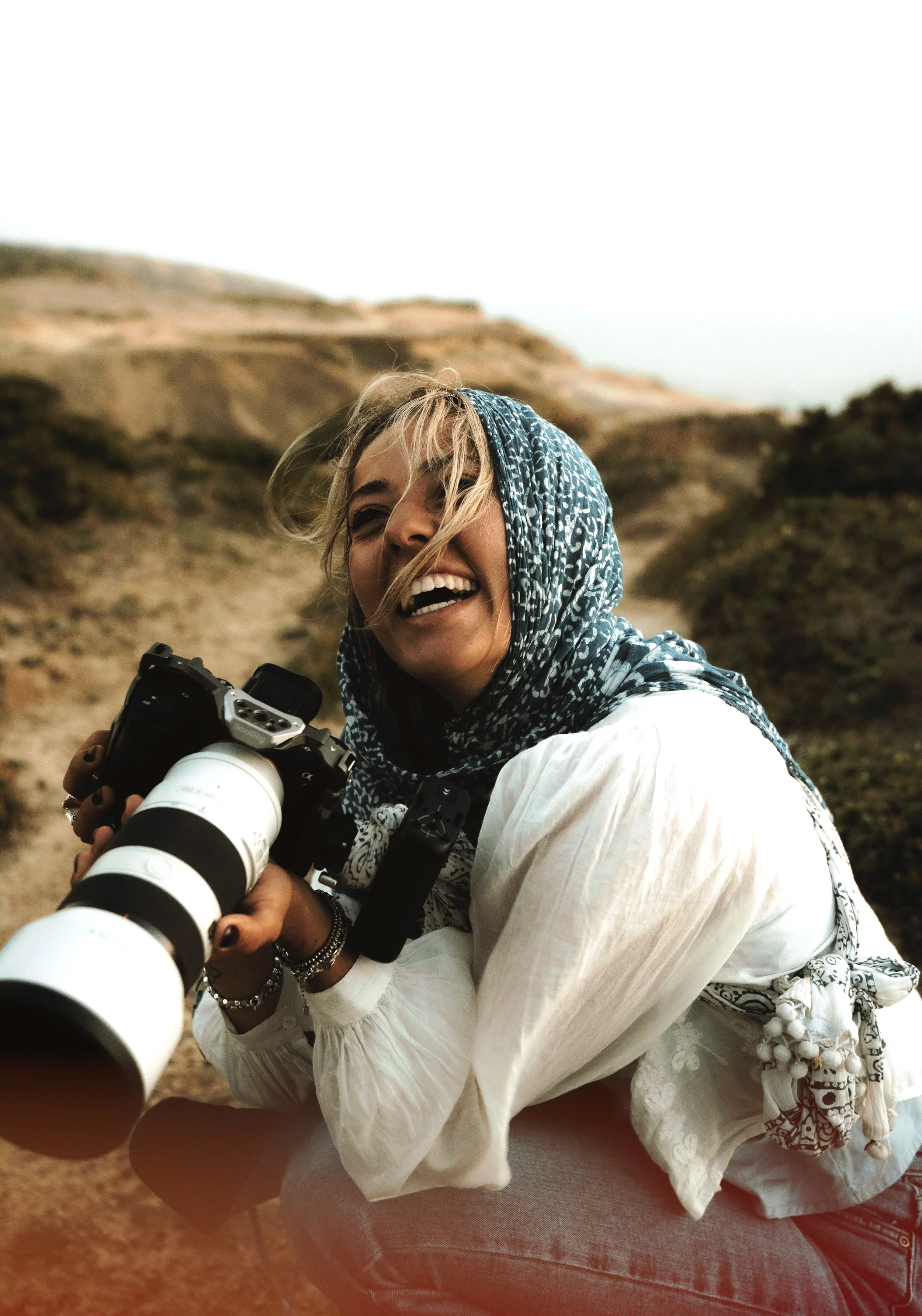 Donna con capelli biondi e sorriso si prende una foto con un grande telescopio astronomico, indossa una sciarpa blu con un motivo bianco e un abito bianco con dettagli ricamati, seduta su una sedia in un paesaggio rurale