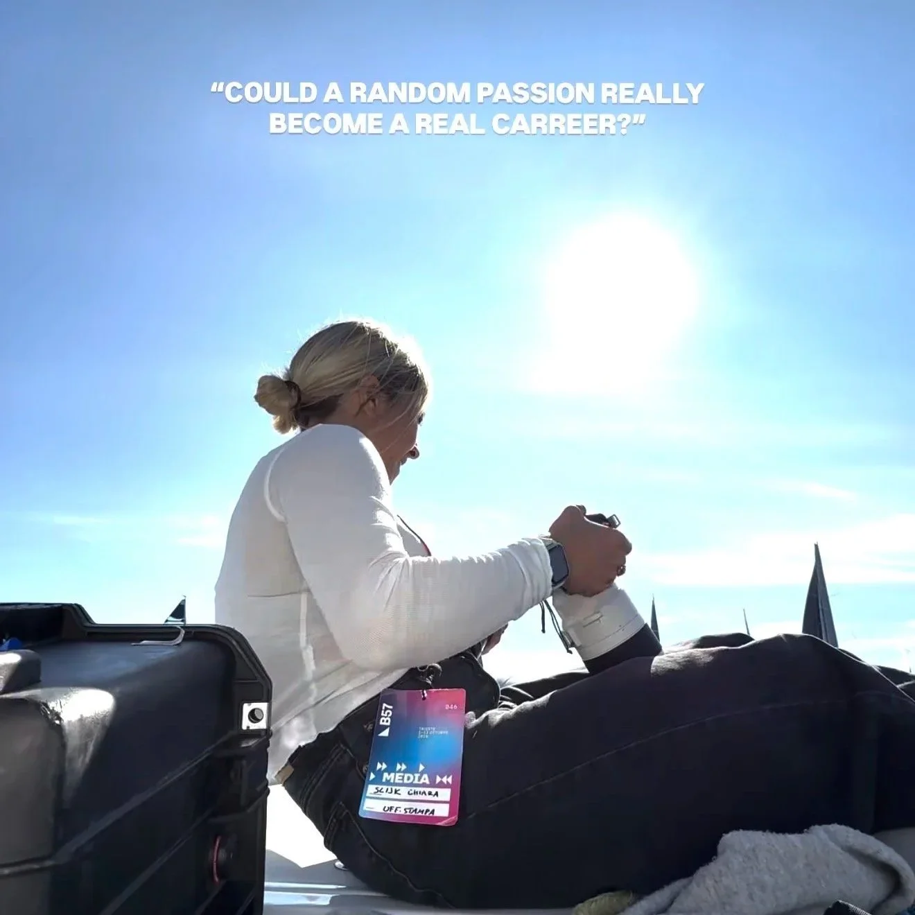 Una donna con i capelli biondi raccolti in uno chignon, vestita di bianco, che scatta una fotografia con una fotocamera reflex, seduta su una superficie con uno sfondo di cielo azzurro e sole splendente.