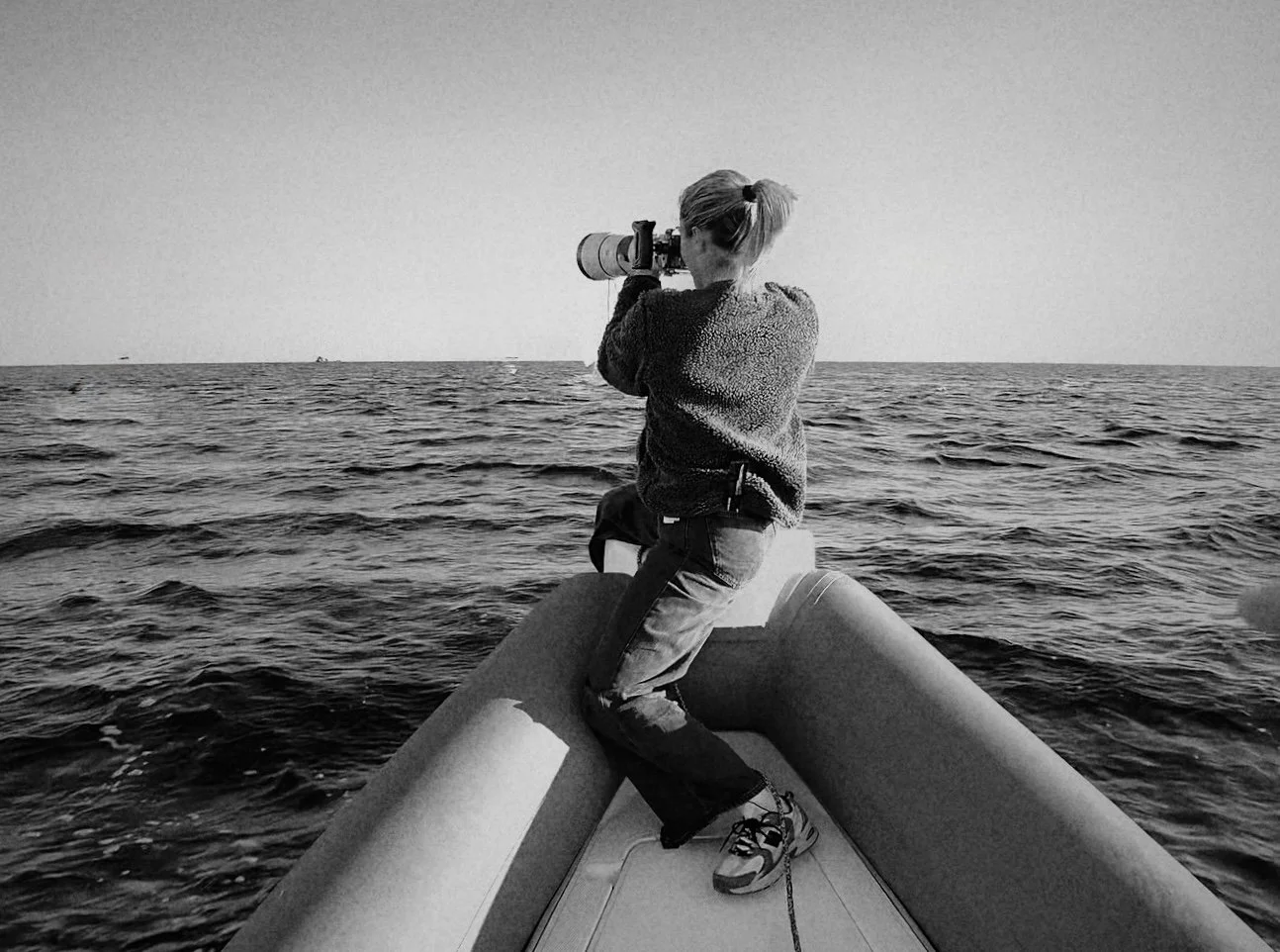 Una donna con cappotto grigio e scarpe sportive, seduta su un'unità di gonfiaggio di una barca, guarda l'orizzonte con un teleobiettivo mentre naviga in mare.