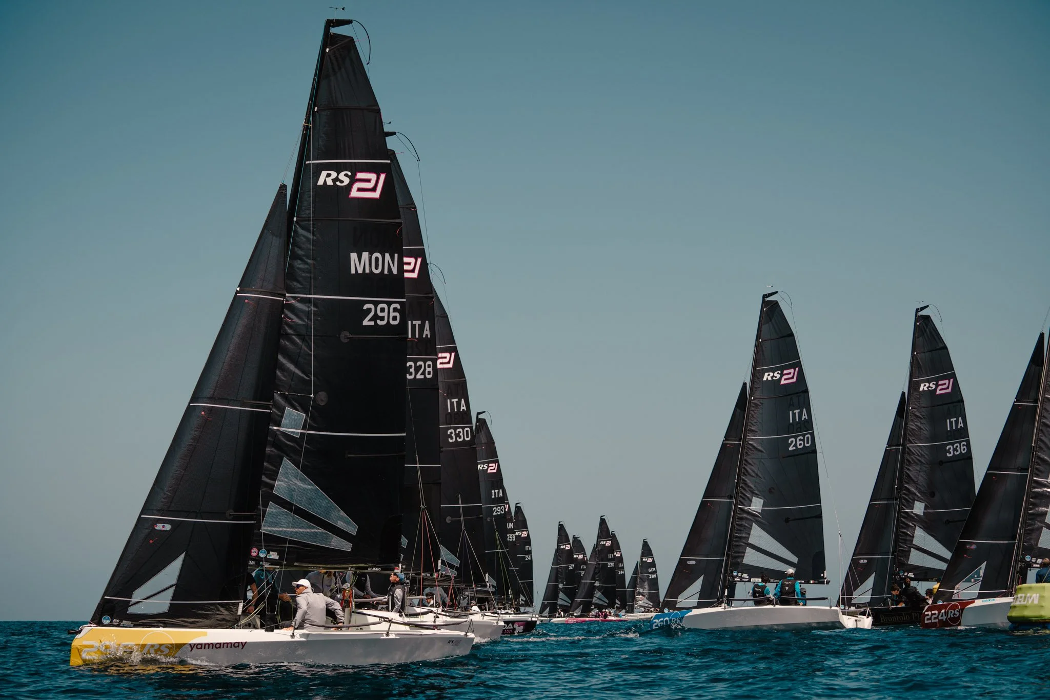 Regata di barche con vele nere e puntinate al largo del mare, con persone a bordo che prevedono la regata in una giornata di cielo sereno.