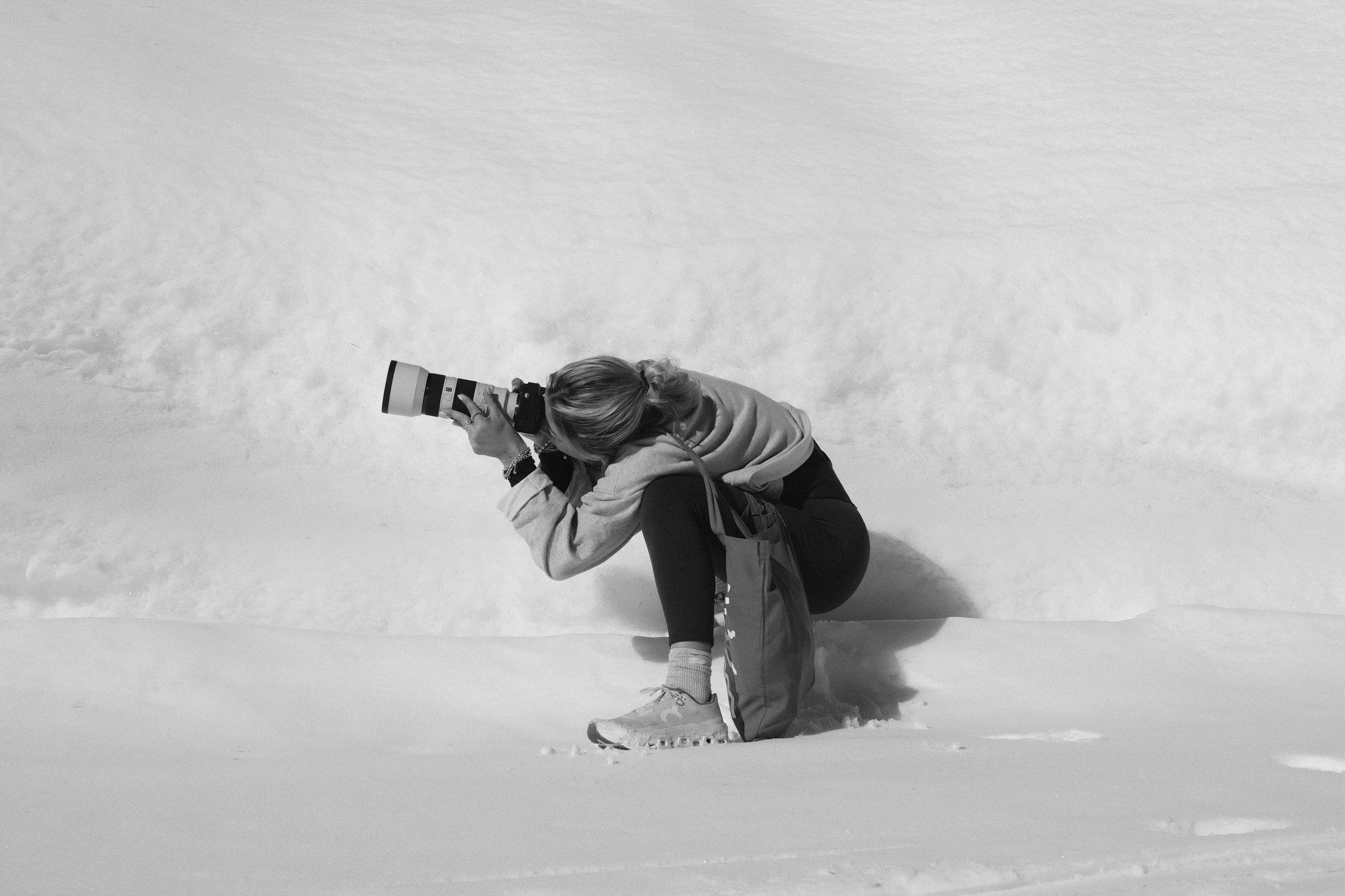 Una persona che scatta una foto sulla neve, si trova in posizione accovacciata, indossa abbigliamento invernale e tiene una fotocamera con un obiettivo teleobiettivo