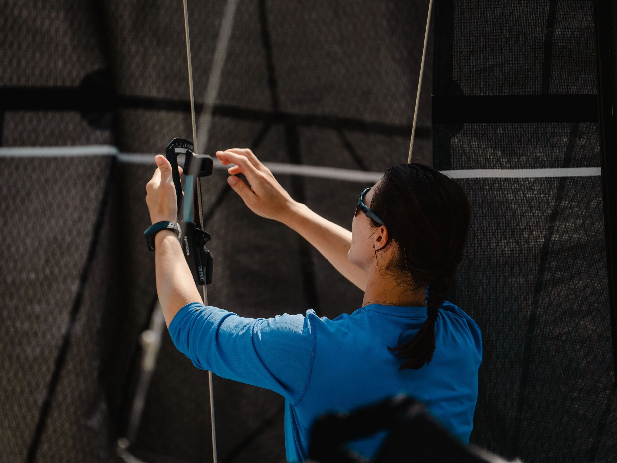 Una donna che indossa occhiali da sole e una maglietta blu sta usando uno strumento di misurazione, mentre è sotto una struttura sospesa con cavi e una rete.