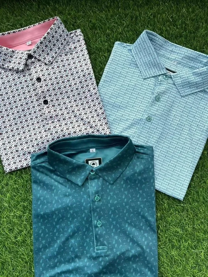 Three folded men's polo shirts with patterned designs, laid out on green grass. The shirts are in different colors and patterns.
