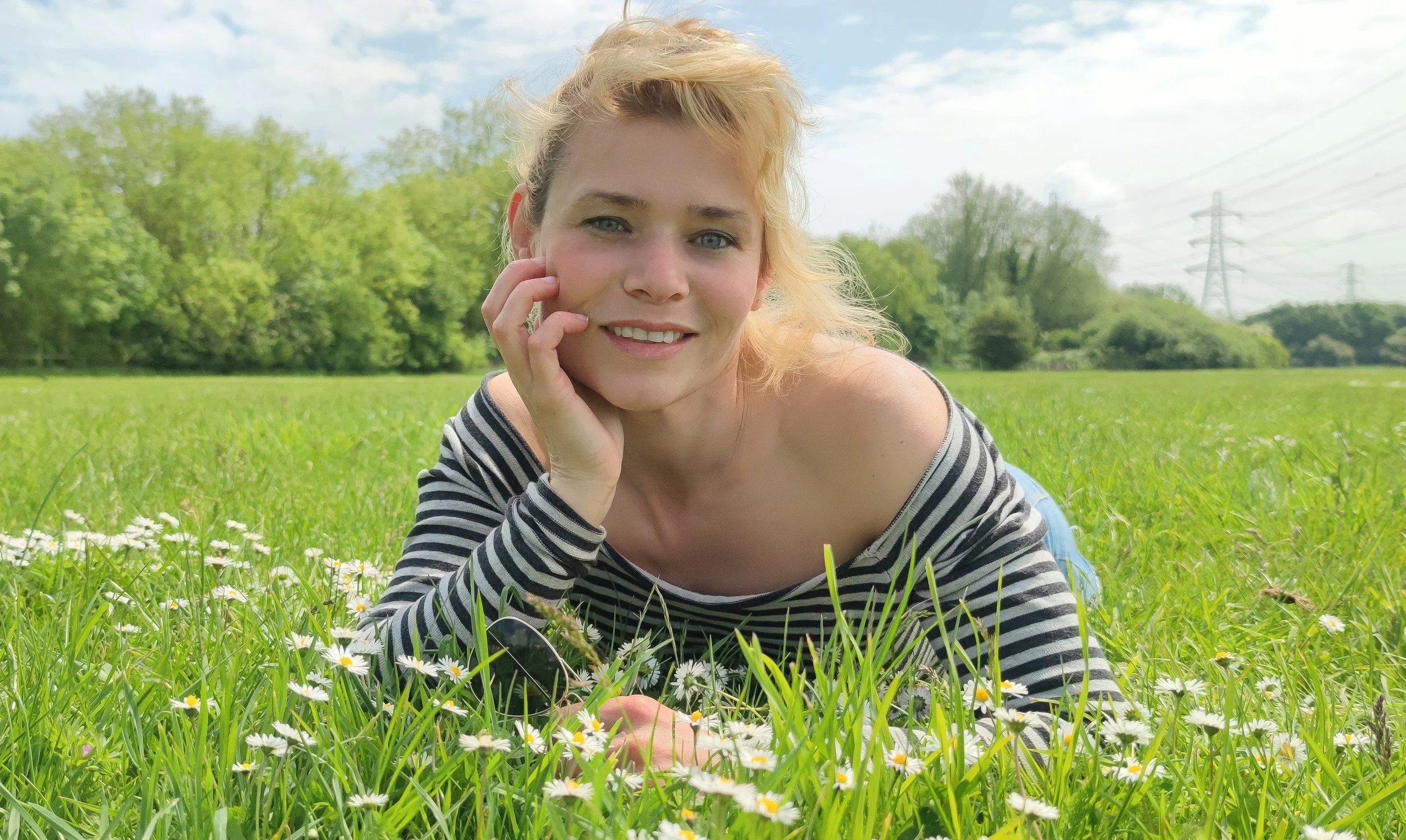 Une jeune femme aux cheveux courts blonds, portant un pull à rayures noires et blanches, repose dans un champ de fleurs blanches avec un sourire, entourée de verdure et sous un ciel partiellement nuageux.
