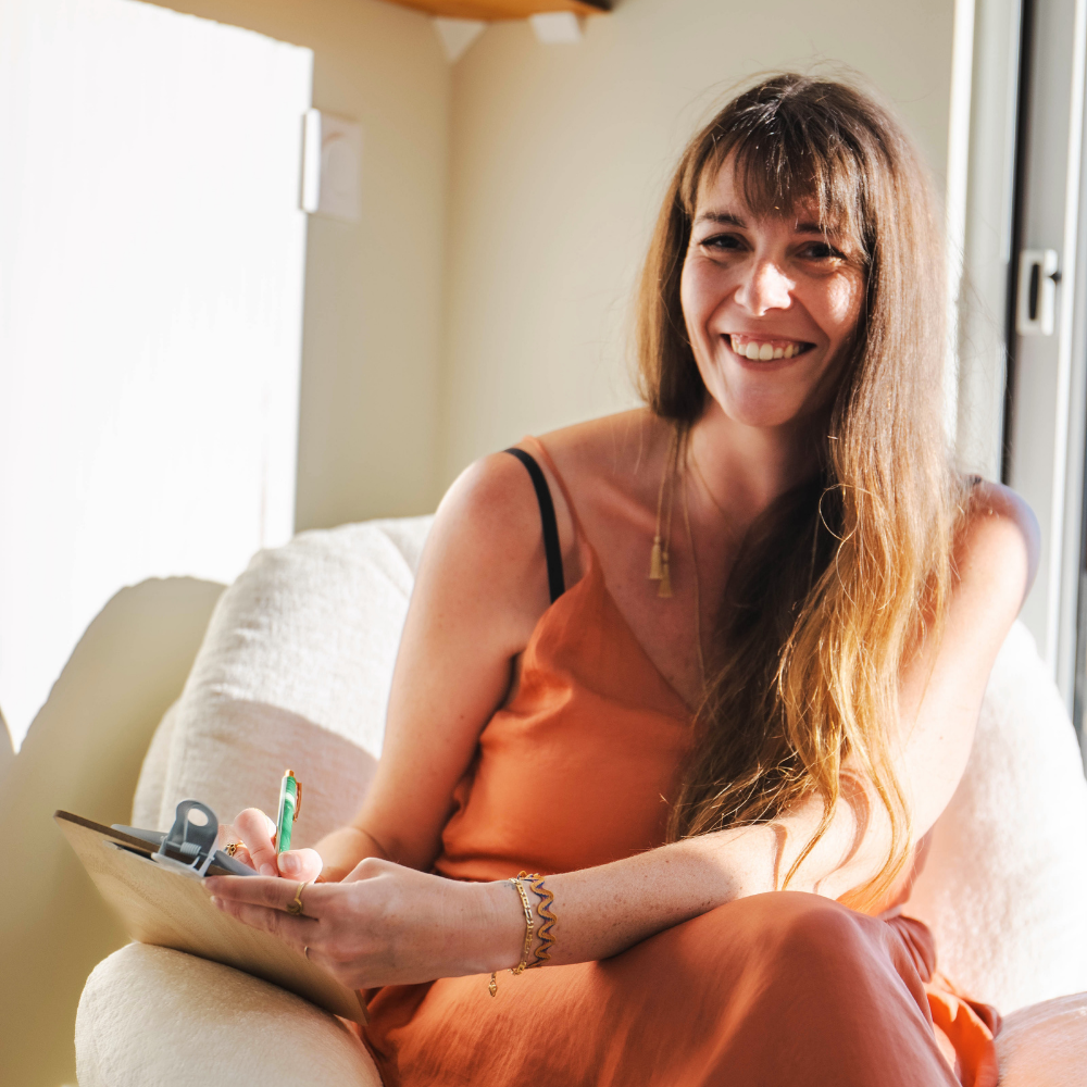 Une femme souriante avec des cheveux longs, assise sur un canapé, en train de tenir un carnet et un stylo, à côté d'une fenêtre à la lumière naturelle.