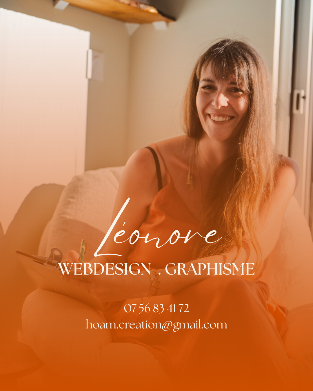 Une femme souriante avec des cheveux longs, assise sur un canapé dans une pièce chaleureuse, portant une robe orange et tenant un carnet.