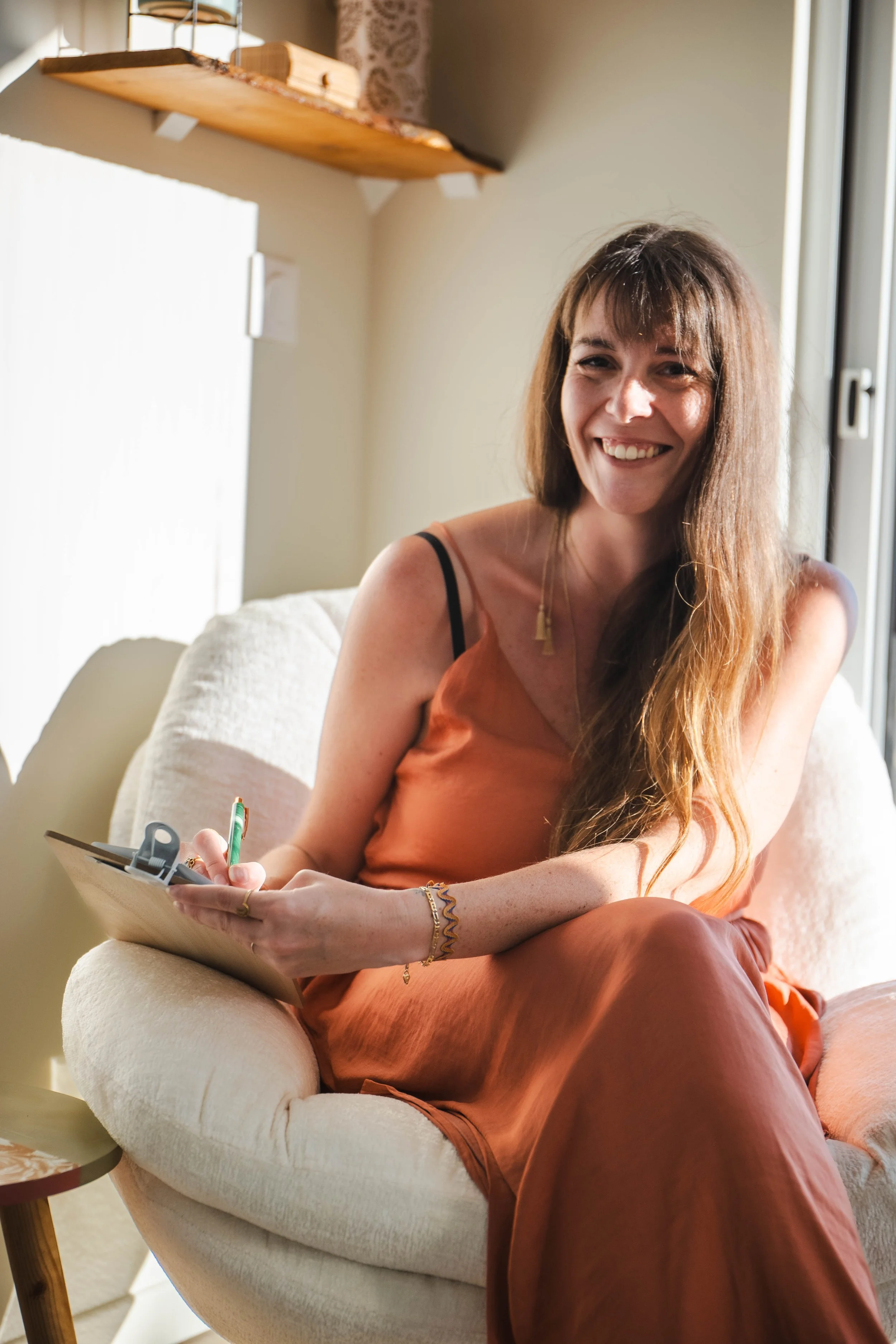 Une femme souriante avec des cheveux longs, assise sur un canapé, en train de tenir un carnet et un stylo, à côté d'une fenêtre à la lumière naturelle.
