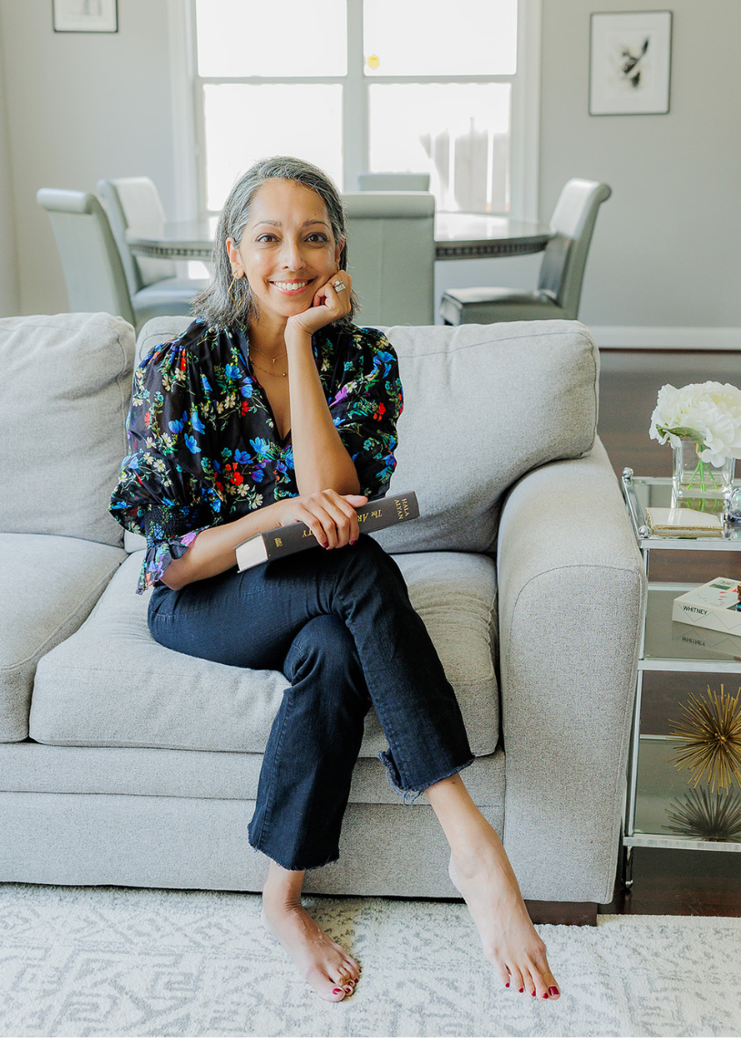 Aditi Pemmaraju, Houston physician relocation realtor, sitting in a bright living room holding a book and smiling.