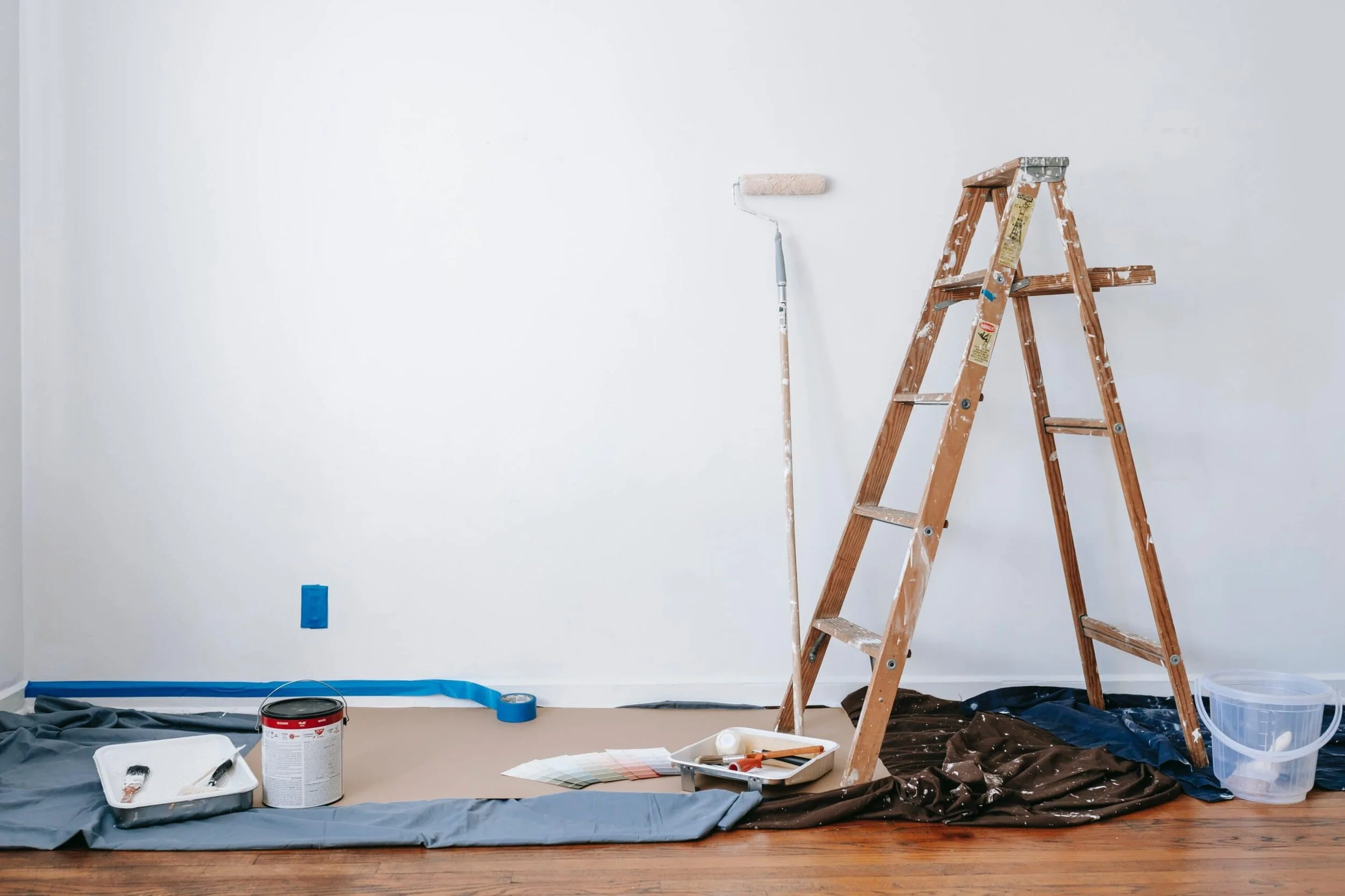 Room being painted with painting supplies including a ladder, paint roller, paint tray, paint can, painter's tape, color swatches, and protective sheet on the floor.