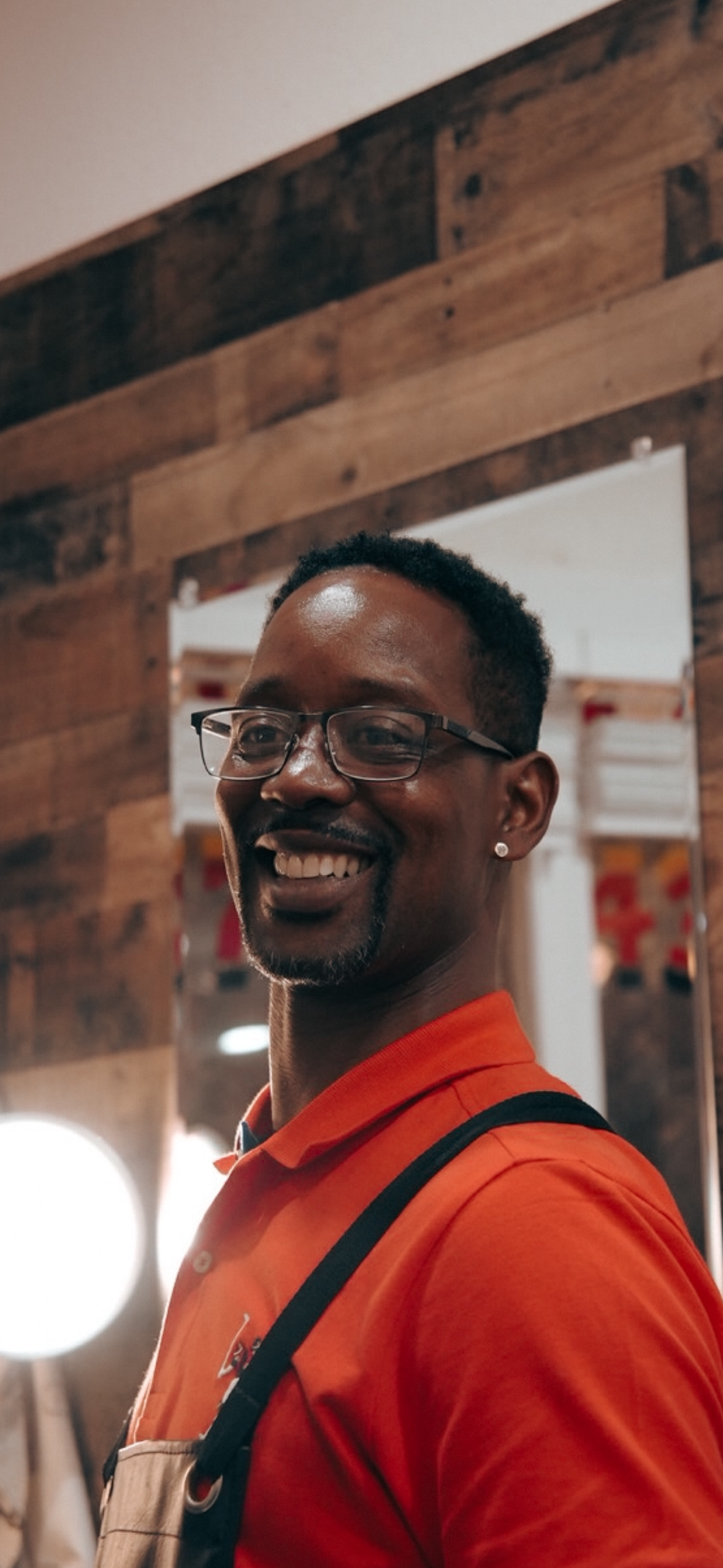 Smiling man wearing glasses and an orange shirt in front of a wooden wall with a mirror.