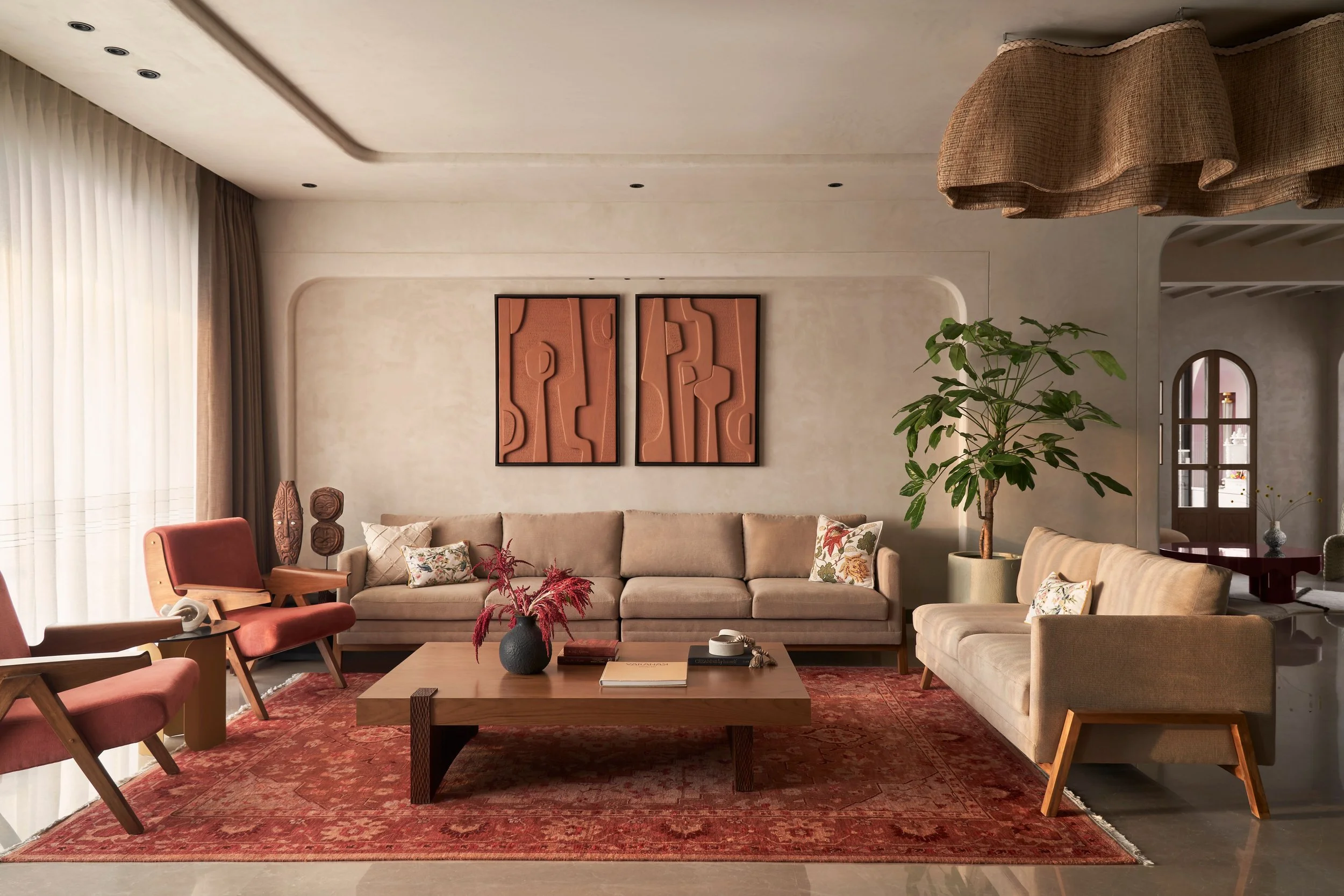 A cozy living room with beige sofas, wooden chairs with pink cushions, wooden coffee table, decorative pillows, wall art, houseplant, and large windows with curtains, on a red patterned rug.