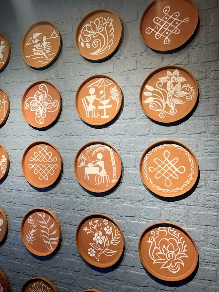 Decorative wall plaques featuring white line art of floral patterns, abstract designs, and scenes of people relaxing and playing music on terracotta-colored plates hanging on a gray brick wall.