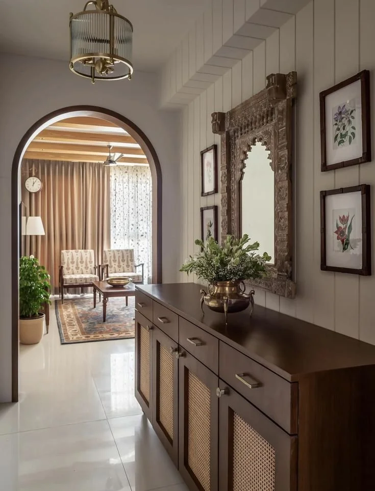 Entryway with a dark wooden sideboard, ornate mirror, and framed artwork on white paneled wall, leading to a sitting area with armchairs, a rug, and curtains.