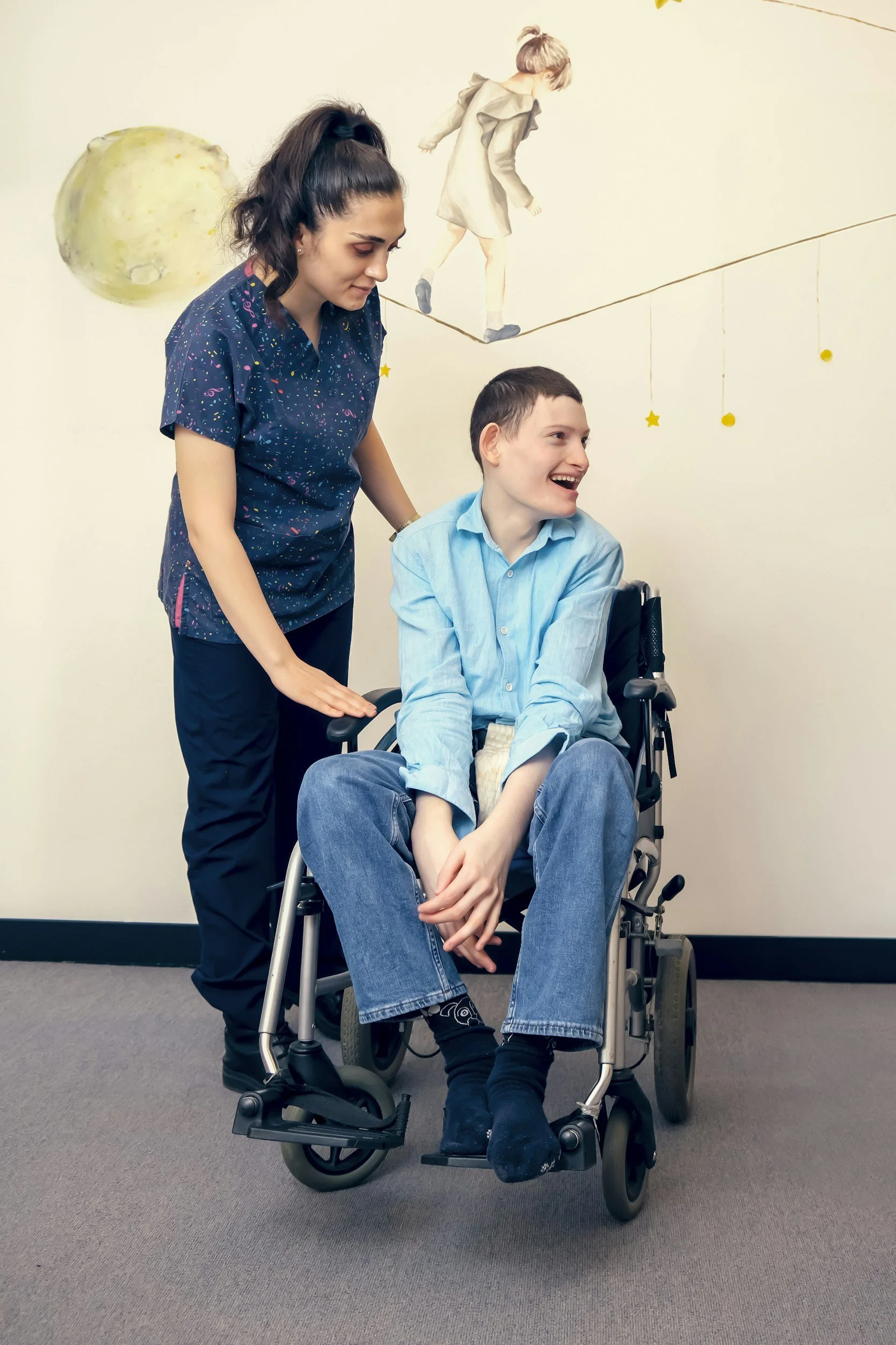 A young man sitting in a wheelchair with a smiling woman standing next to him, holding his shoulder, in front of a wall with a mural of a girl walking on a tightrope. The young man is wearing a light blue shirt and jeans, and the woman is wearing a dark blue shirt with colorful specks and black pants.