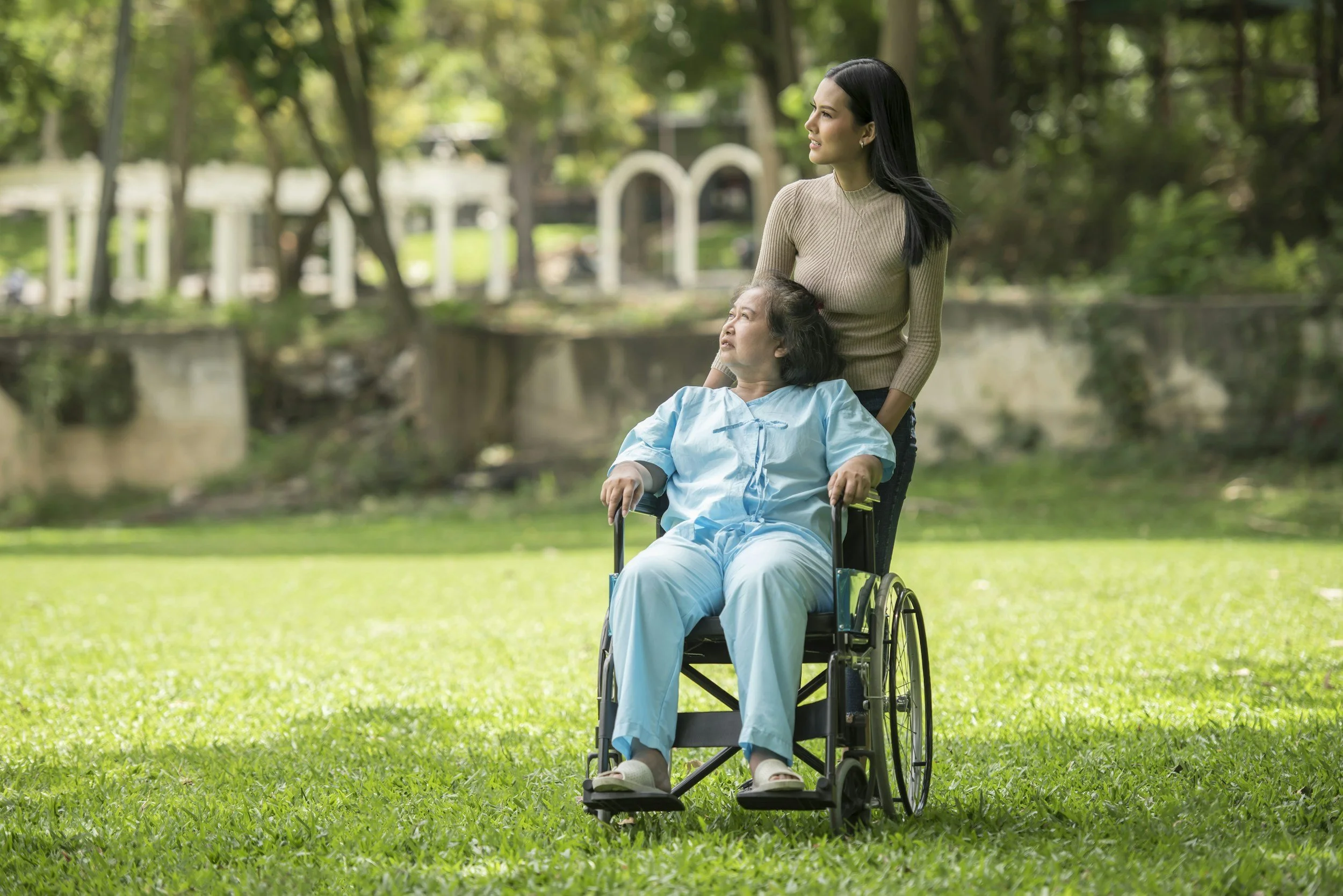 A woman with dark hair standing behind a woman in a wheelchair that is outdoors on a grassy area, with trees and a bridge in the background.