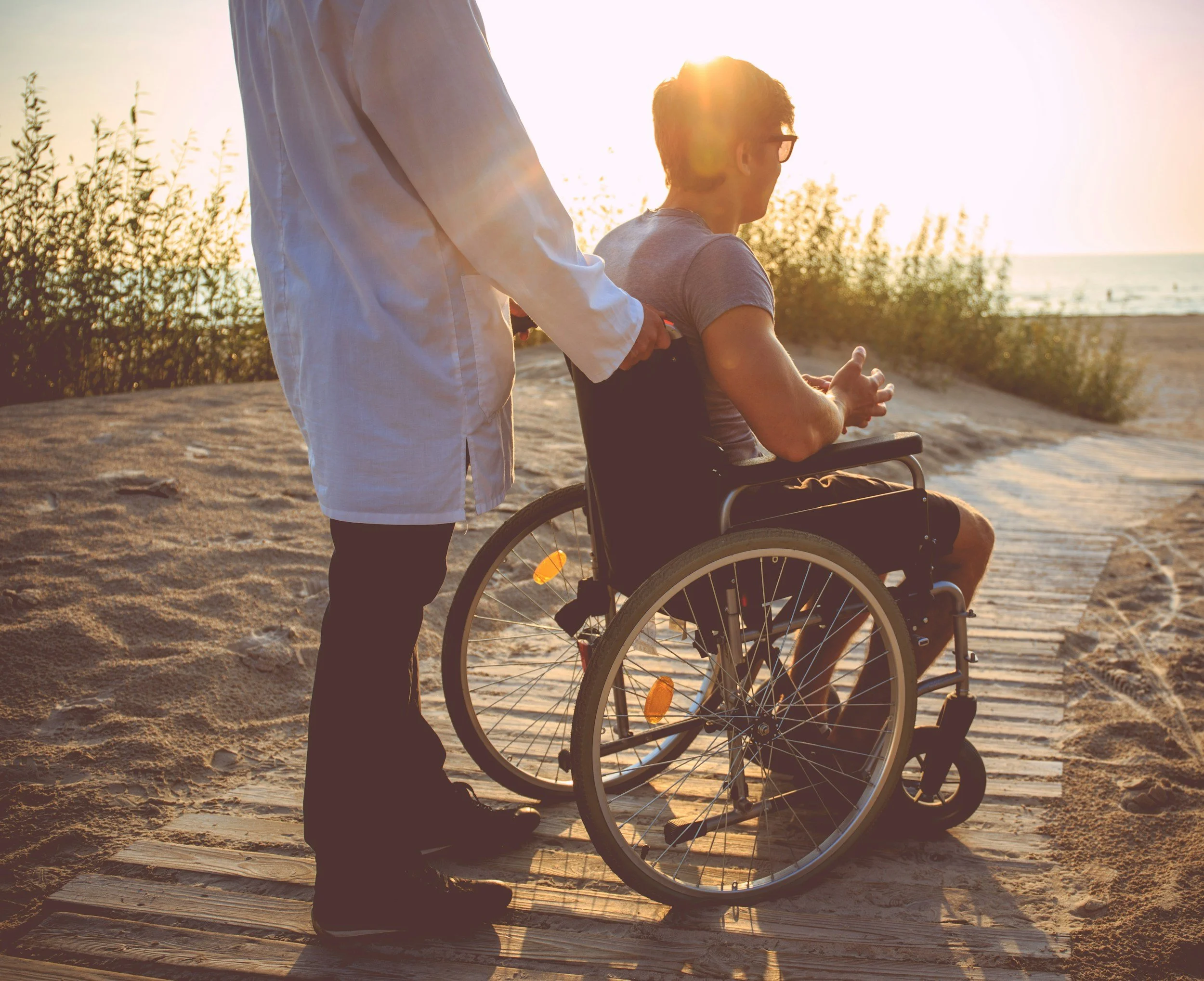 A person in a wheelchair on a sandy beach during sunset, with another person standing nearby and holding a device. The sunlight creates a warm glow, and there are some bushes and the ocean in the background.