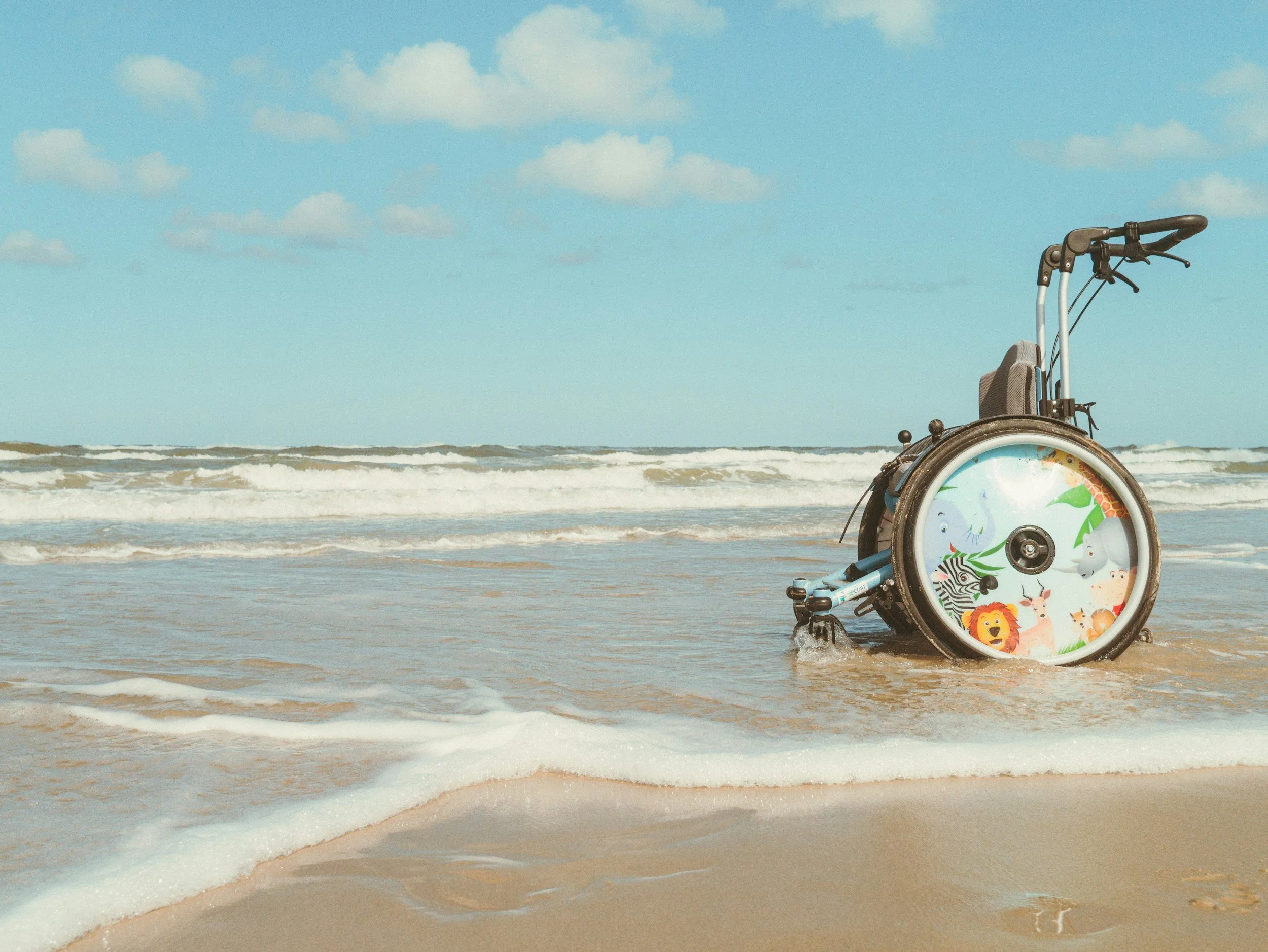 A wheelchair with a colorful animal-based design on the wheel, positioned on a sandy beach with foamy waves and a clear sky with some clouds.