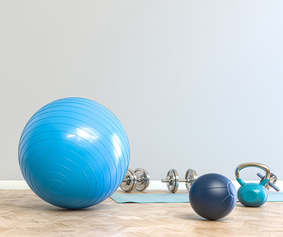 Home gym setup with large blue exercise ball, smaller dark blue exercise ball, kettlebell, dumbbells, and a yoga mat on wooden floor against plain wall.