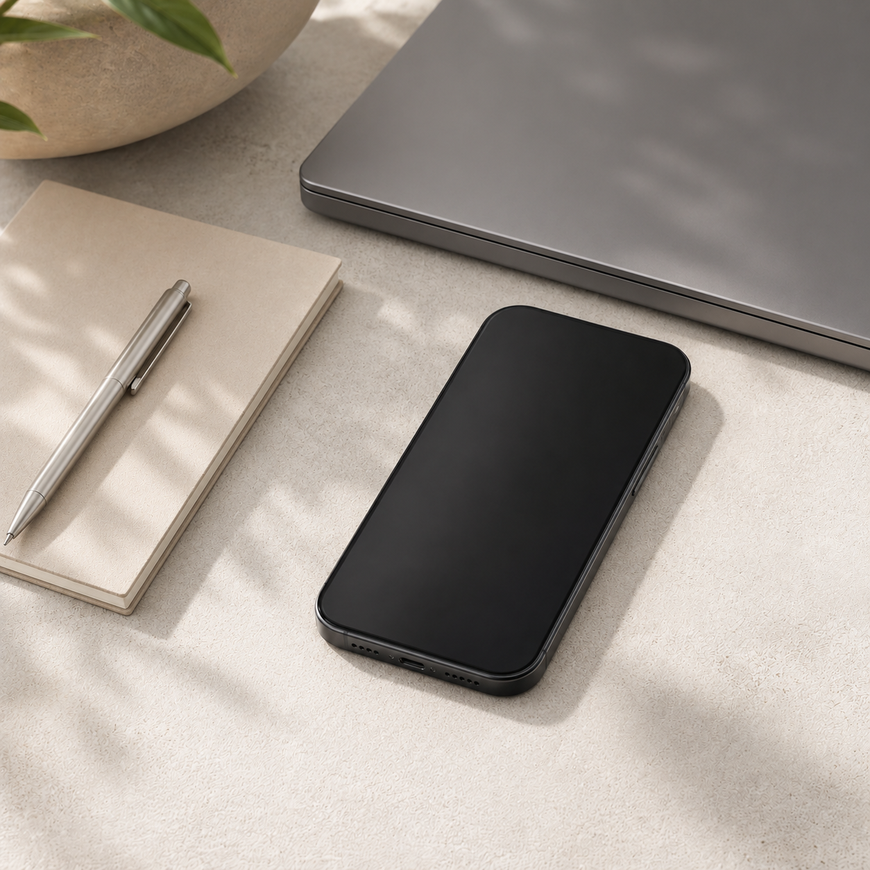 A modern workspace with a black smartphone, a silver laptop, a closed beige notebook, and a silver pen on a light-colored surface.