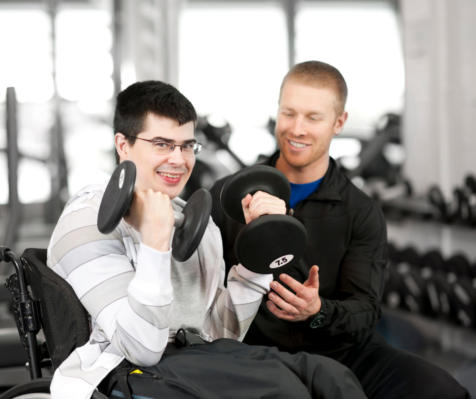 A man in a wheelchair lifting dumbbells while a trainer or therapist assists and encourages him in a gym or fitness center.