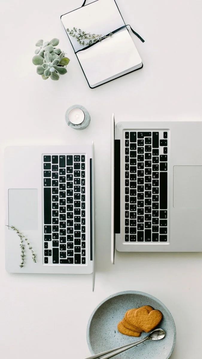 Top-down view of a minimalist workspace with a laptop, external keyboard, notebook, succulent, candle, stylus, and plate of cookies